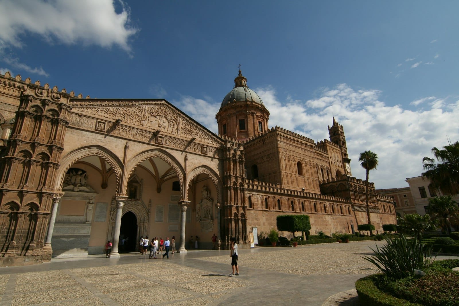 Weite Ansicht der Kathedrale von Palermo in Sizilien mit ihren arabisch-normannischen Architekturmerkmalen, mit Bögen, Steinschnitzereien, Kuppel und Menschen draußen.