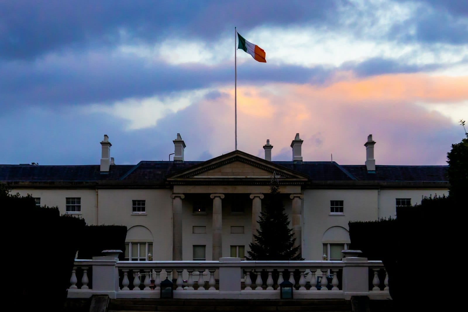 Vista frontale di Áras an Uachtaráin, residenza presidenziale irlandese, con la bandiera irlandese al vento, colonne bianche e un cielo serale spettacolare.