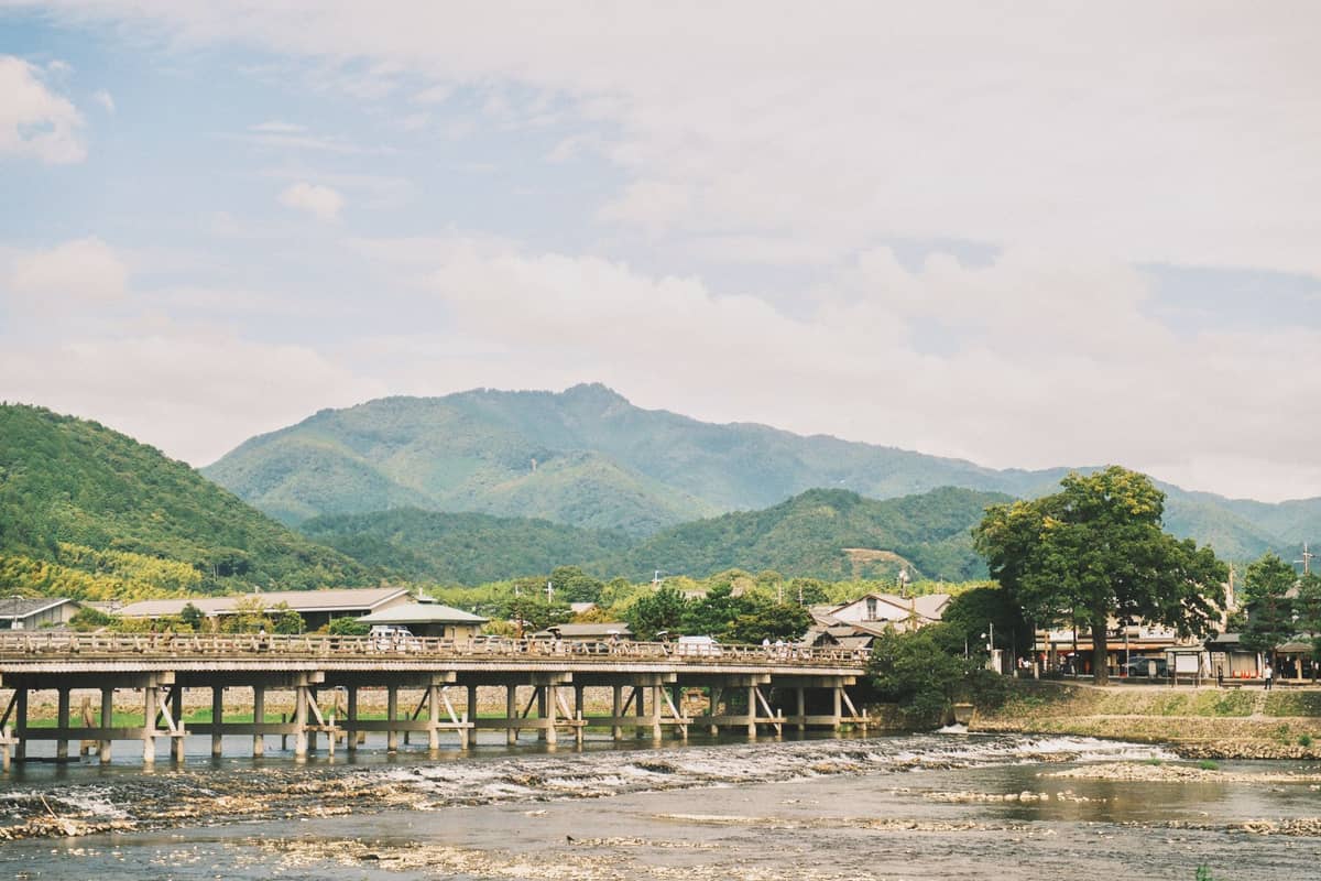 Scenic view of Arashiyama’s Togetsukyo Bridge spanning the Katsura River with lush mountains and village buildings in the background on a sunny day.