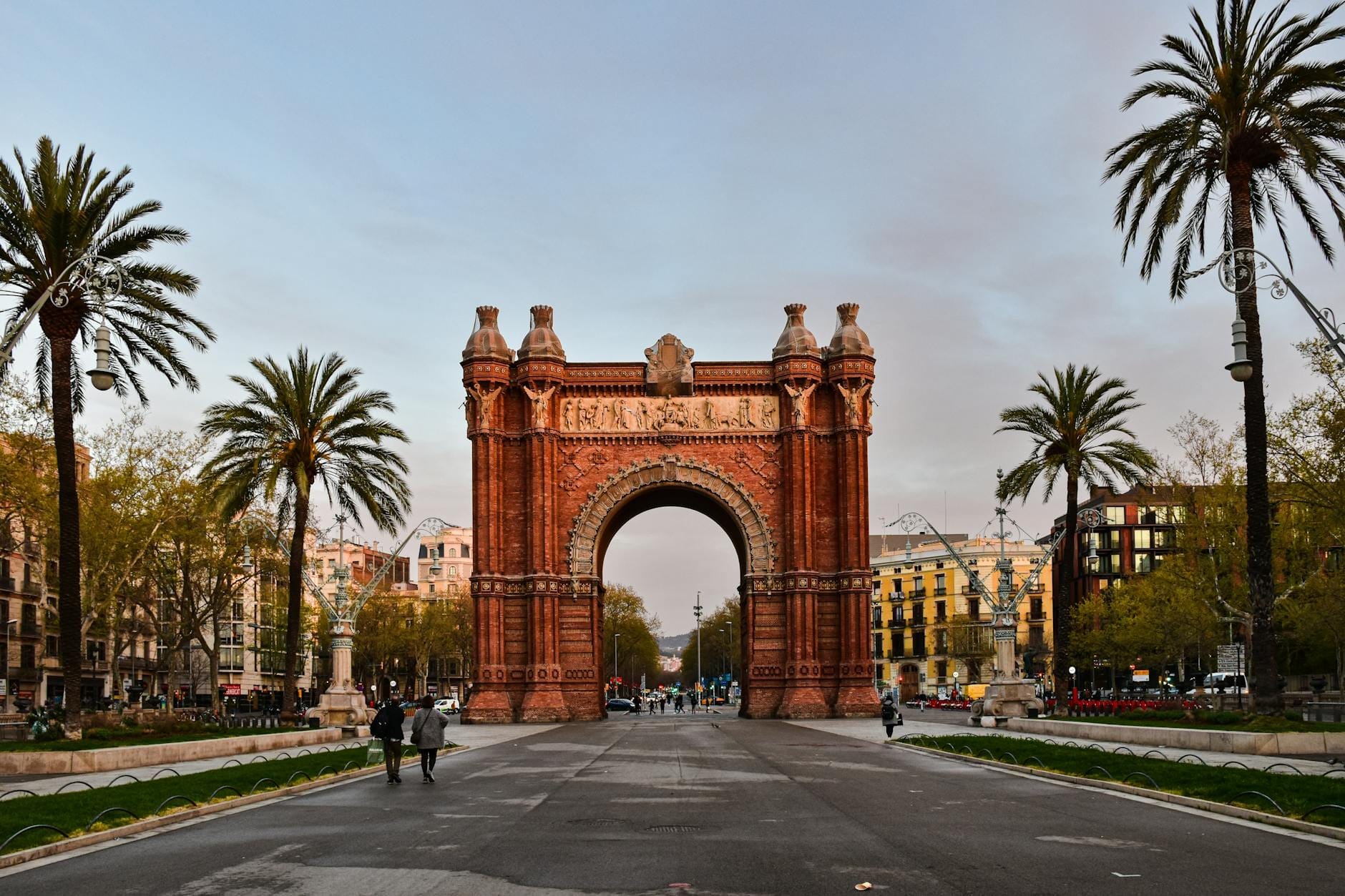 Wide view of Arc de Triomf in Barcelona with palm trees lining a central promenade. People are walking toward and around the monument under a soft, golden sky.