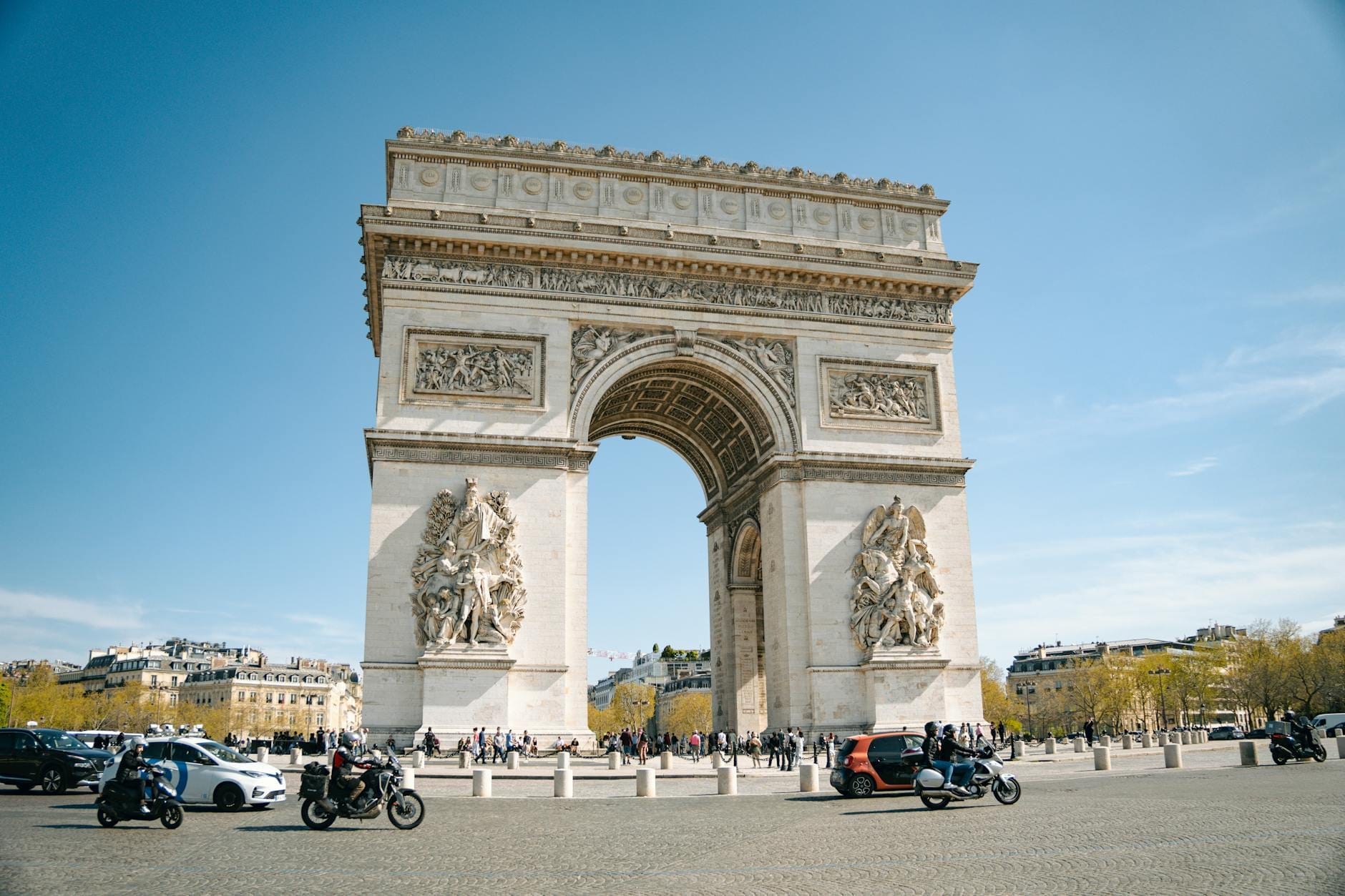 Vista grandangolare dell'Arco di Trionfo di giorno, con cielo azzurro, persone e veicoli intorno al monumento. Sullo sfondo si scorgono edifici parigini e alberi.