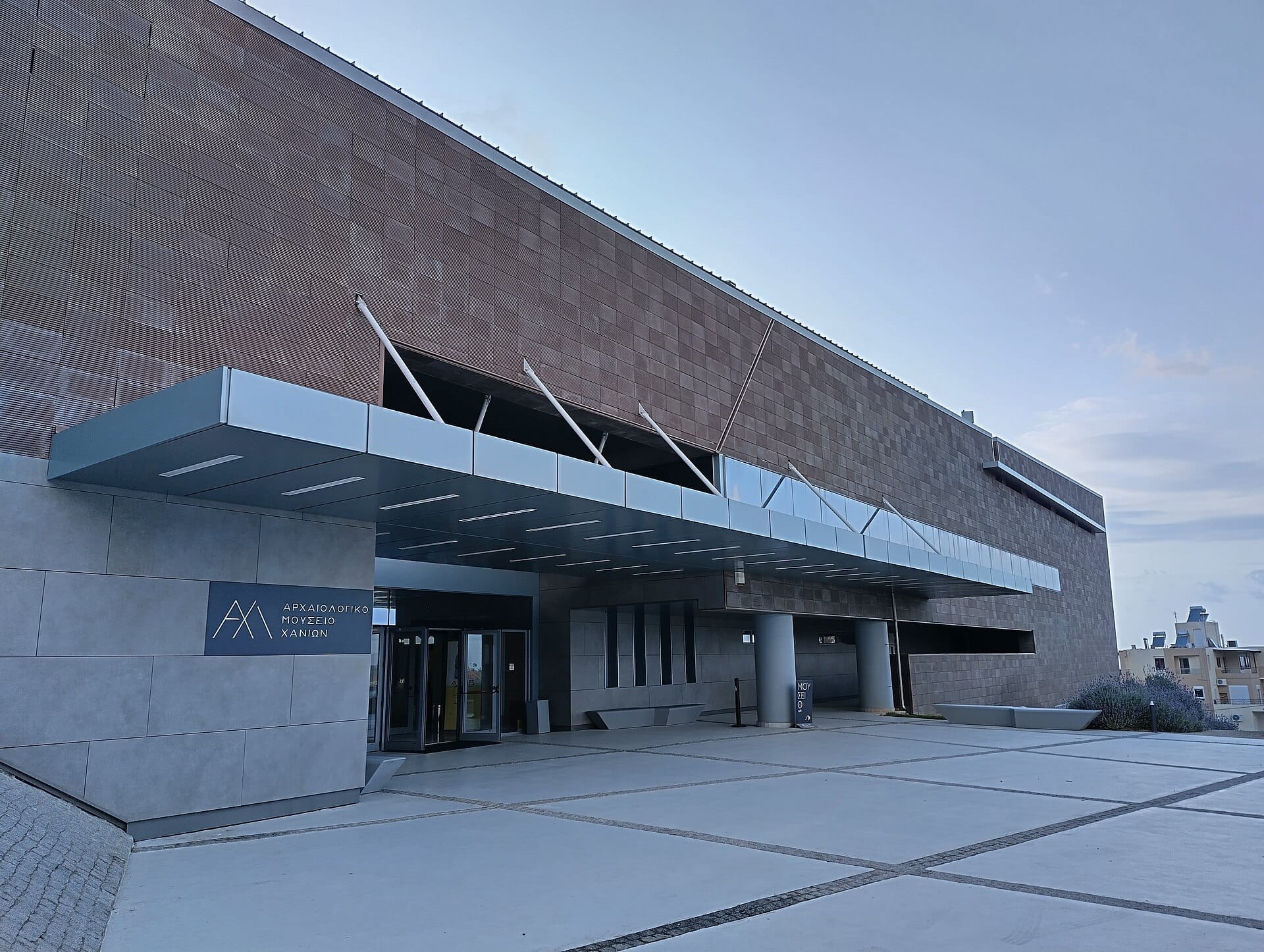 Esterno moderno del Museo Archeologico di Chania, con linee geometriche, grande pensilina in vetro e facciata in mattoni testurizzati nella luce soffusa del giorno.