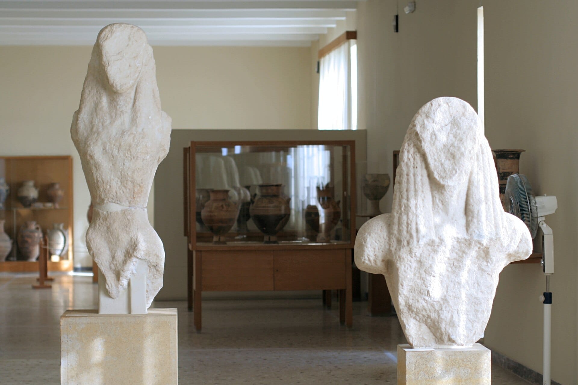 รูปปั้นหินอ่อนโบราณสองชิ้นจัดแสดงบนแท่นในหอแสดงงานที่สว่างสดใสของ Archaeological Museum of Thera พร้อมแจกันดินเผาในตู้กระจกด้านหลัง