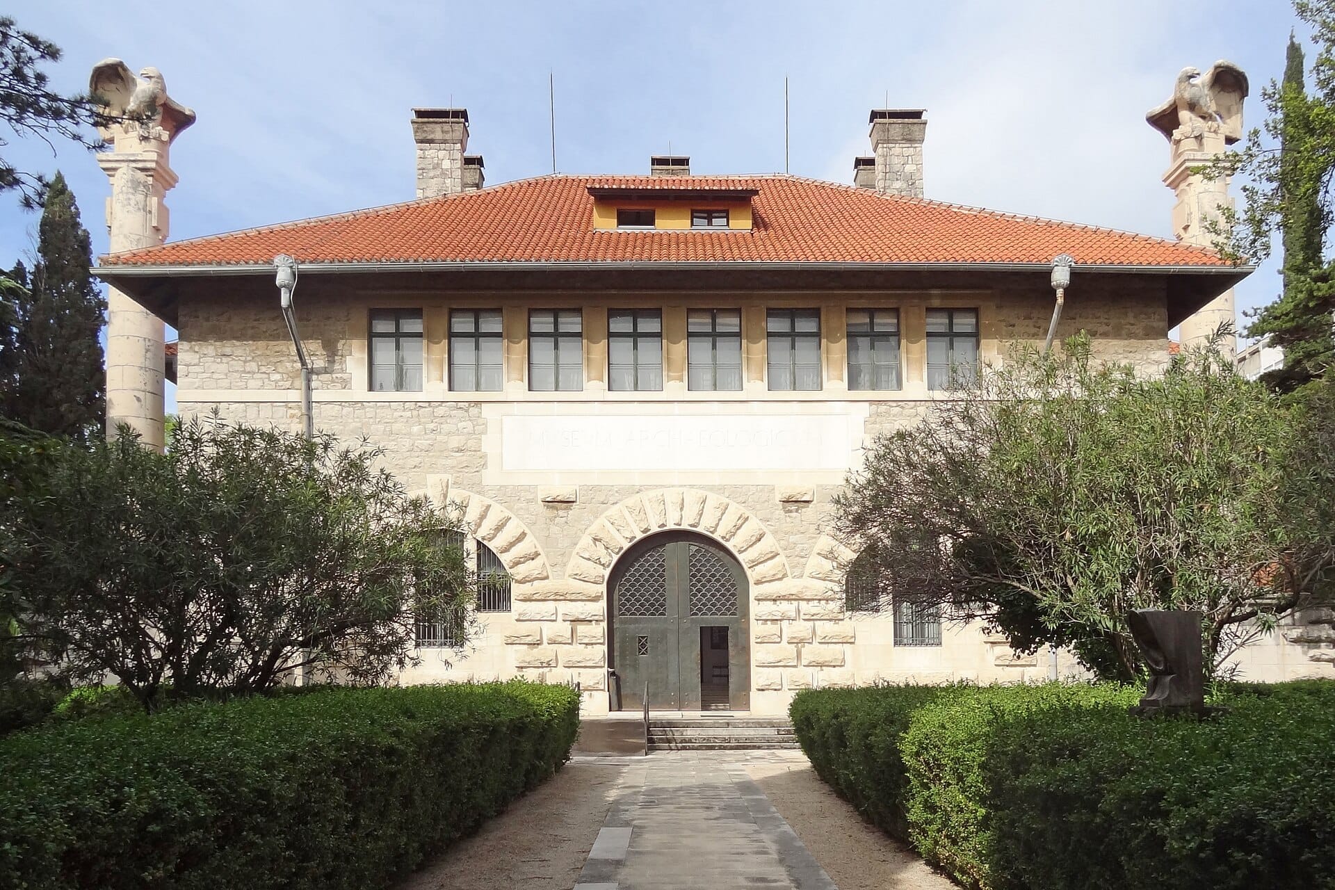 ด้านหน้าของ Archaeological Museum Split ที่มีกำแพงหิน หลังคากระเบื้องสีแดง ทางเข้าโค้งรูปซุ้ม และต้นไม้สมมาตรใต้ท้องฟ้าสดใส