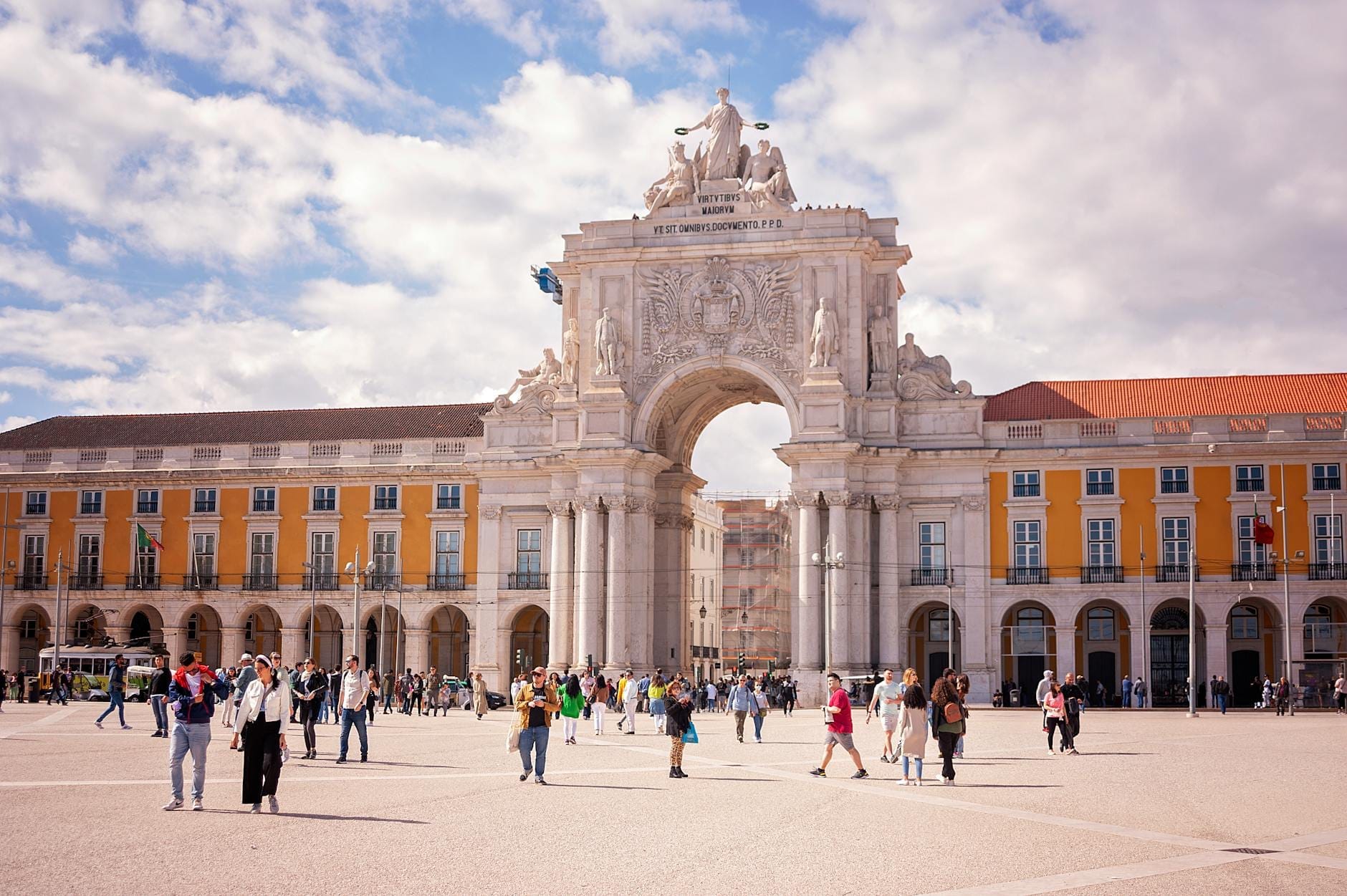 Vista vivace e luminosa dell'Arco da Rua Augusta con la folla di turisti nella Praça do Comércio, edifici gialli e un cielo azzurro con qualche nuvola.