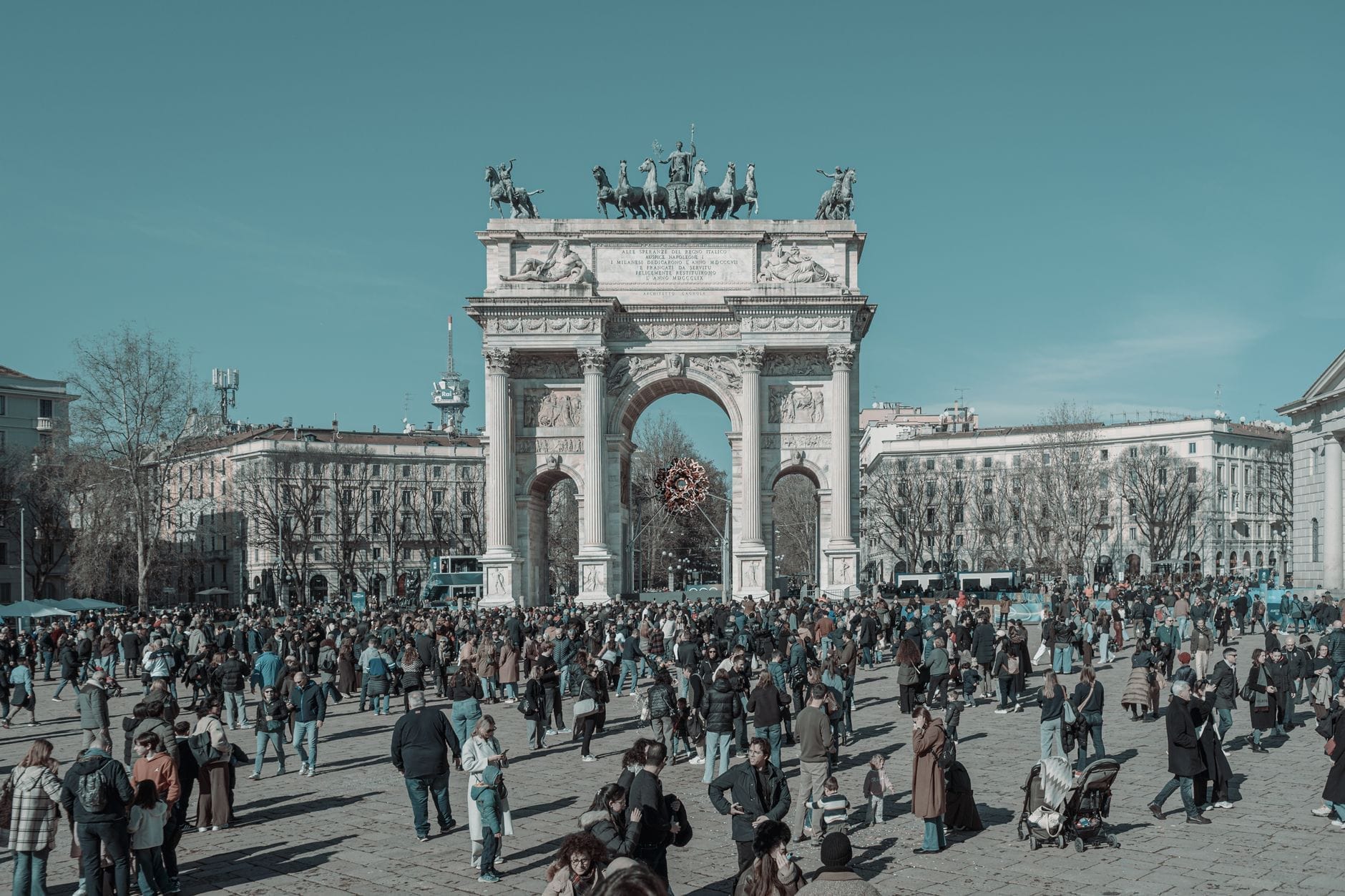 Folla di persone radunata davanti all'Arco della Pace a Milano in una giornata limpida, con l'arco nella sua interezza e la piazza animata.