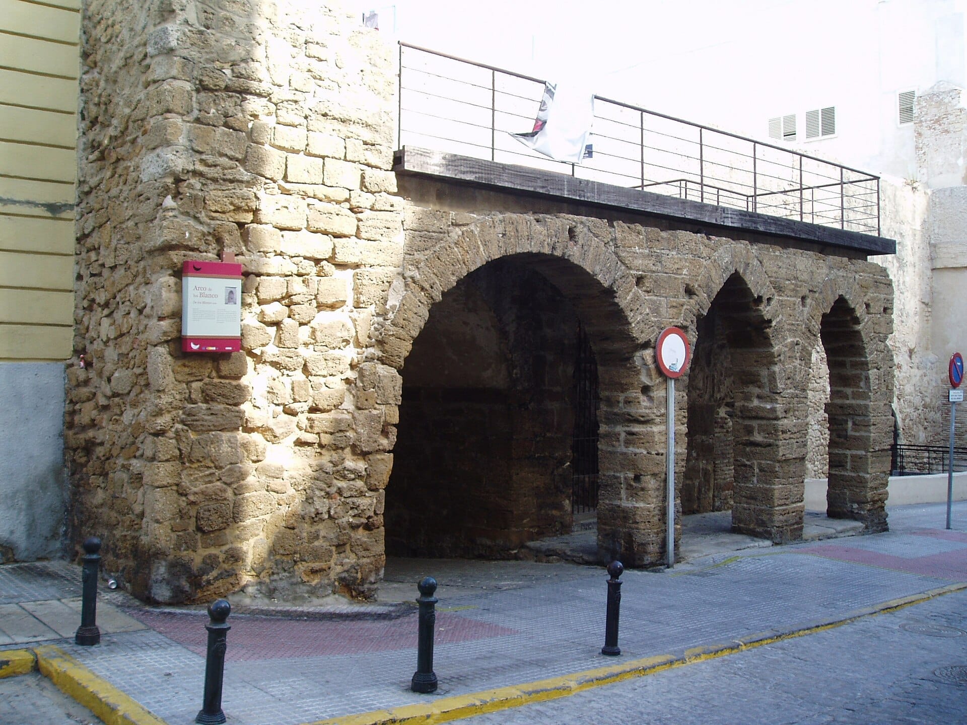 Archi in pietra dell'Arco del Pópulo, Cádiz, con mura medievali consumate e segnali stradali moderni sotto una luce diurna soffusa.