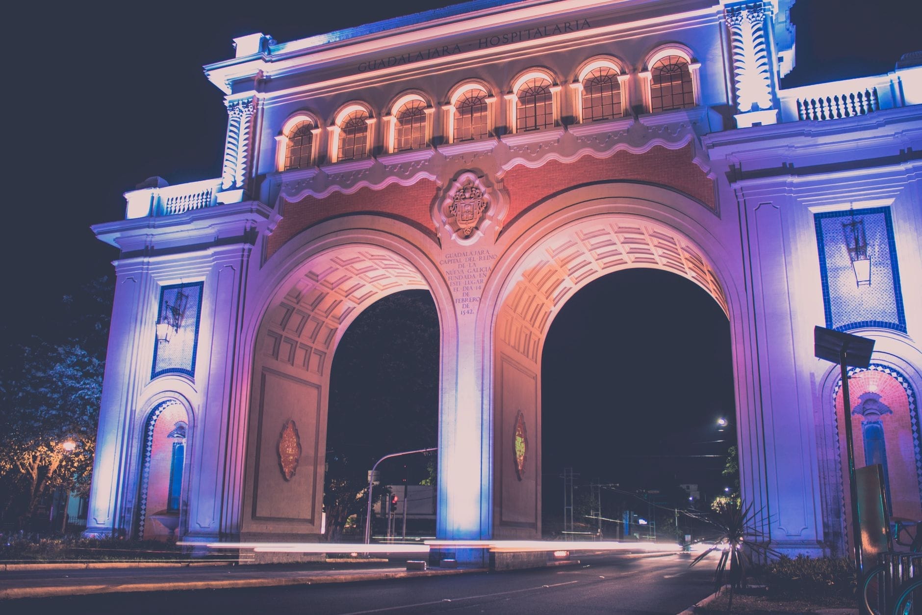 Vista notturna del monumento Arcos de Guadalajara illuminato con luci viola e blu, con entrambi i grandi archi visibili su una strada tranquilla e deserta.