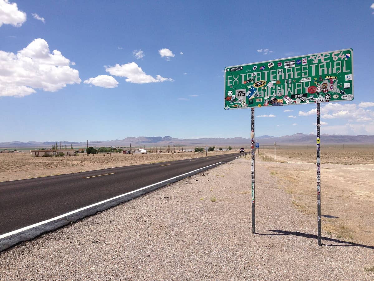 Rambu jalan Extraterrestrial Highway yang tertutup stiker di sepanjang Nevada State Route 375, dengan lanskap gurun yang luas dan pegunungan jauh di bawah langit biru.