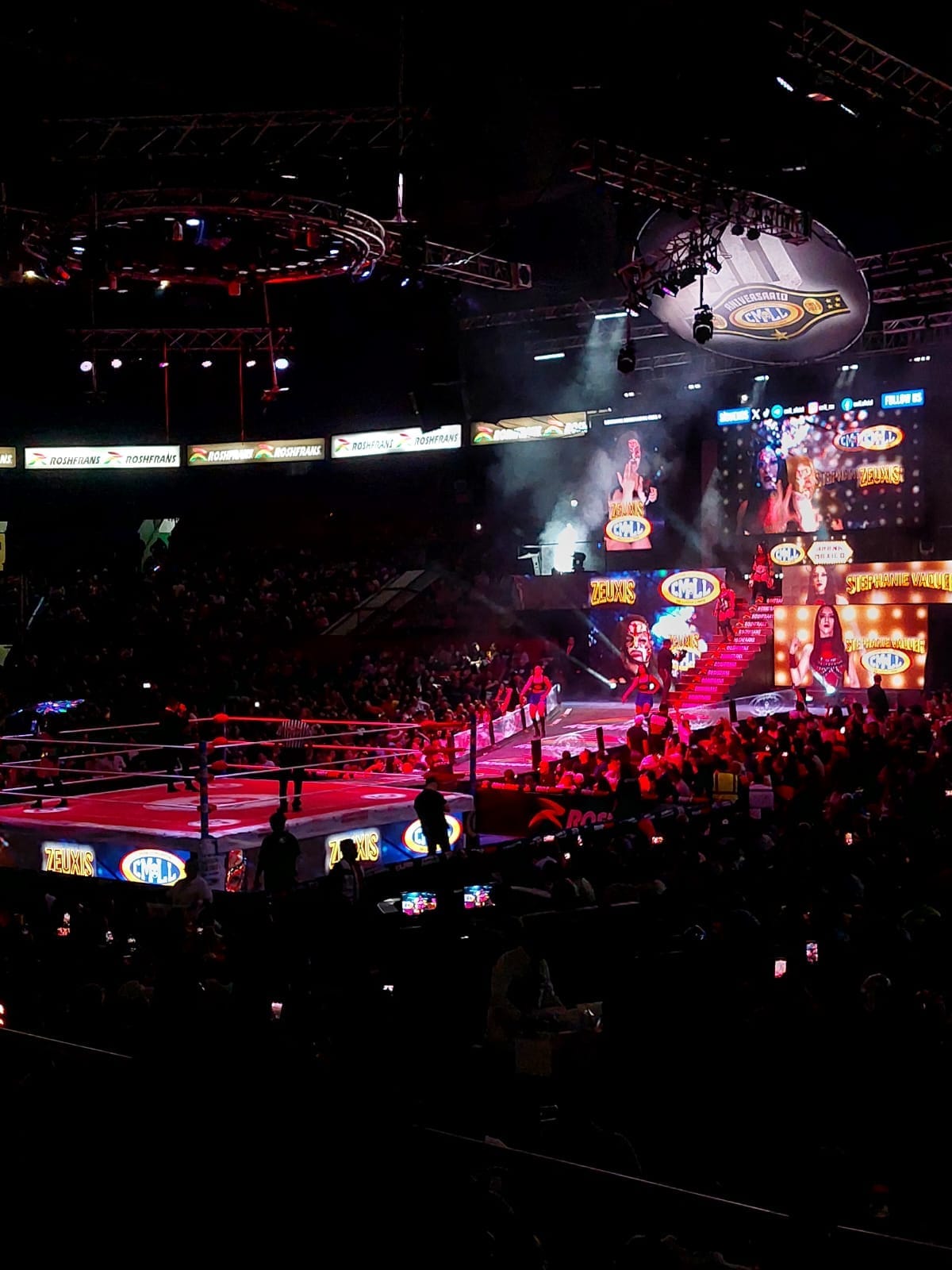 I lottatori entrano nell'illuminato ring dell'Arena México mentre una folla festante riempie l'arena in penombra, con schermi vivaci e faretti dall'alto.