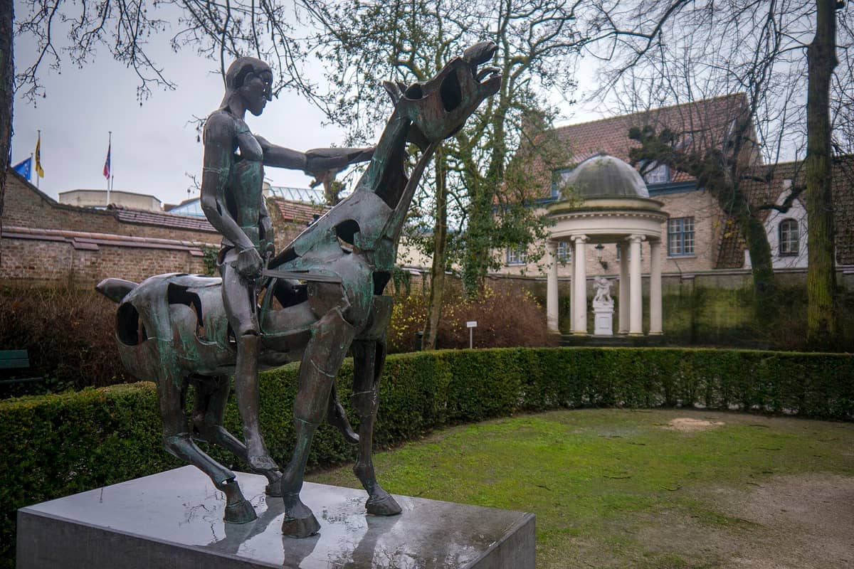 Bronze abstract statue of a rider on horseback in Hof Arents park, with trimmed hedges and a classical pavilion in Bruges, Belgium.