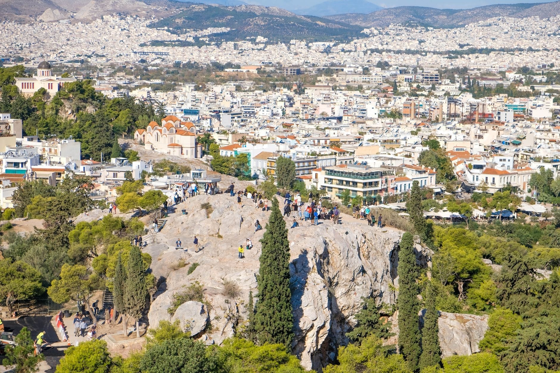 Una veduta soleggiata della Collina dell'Areopago con persone sullo sperone roccioso, circondata da alberi e dal panorama di Atene sullo sfondo.