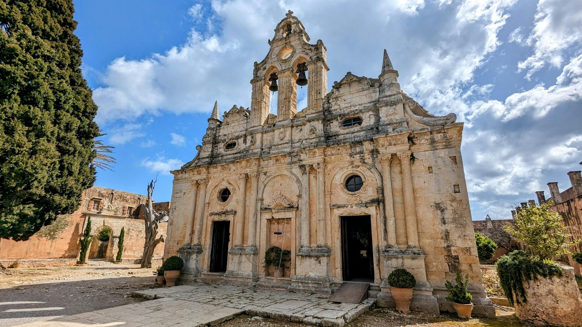 La facciata in pietra riccamente decorata del Monastero di Arkadi a Creta, incorniciata da cipressi e cieli drammatici, con dettagli architettonici e un forte carattere storico.