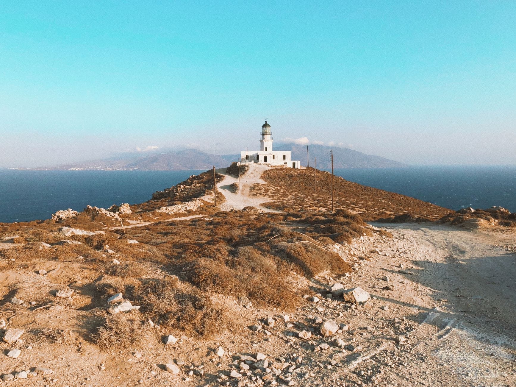 El Faro de Armenistis sobre una colina rocosa con vistas al mar Egeo azul, con un sendero sinuoso que sube hasta él y montañas lejanas al fondo.