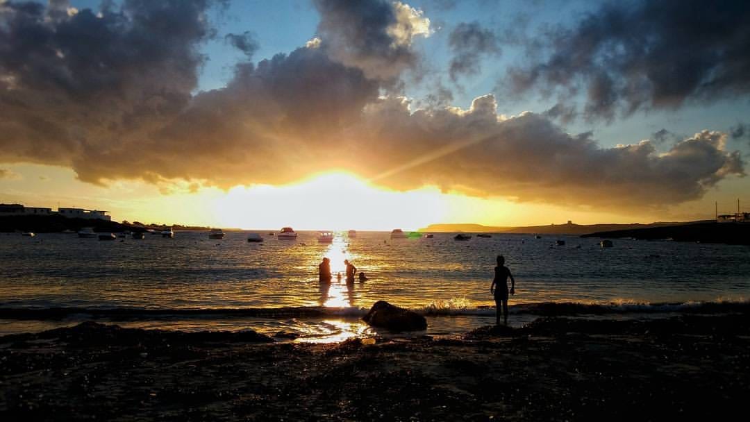 Vista del tramonto ad Armier Bay, Malta, con sagome di persone sulla spiaggia, barche su acque calme e nuvole spettacolari nel cielo.