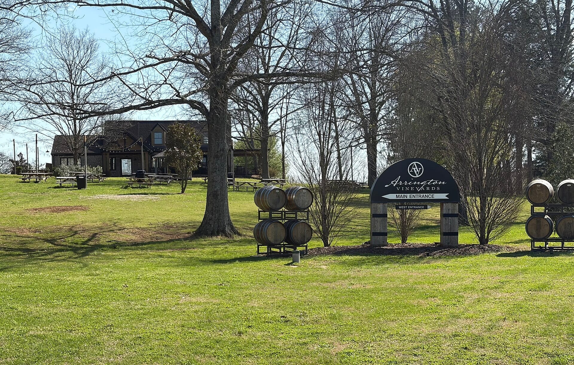 Letrero de la entrada principal de Arrington Vineyards, barriles de vino y un edificio rústico situado en un terreno con hierba y árboles bajo la luz del día.