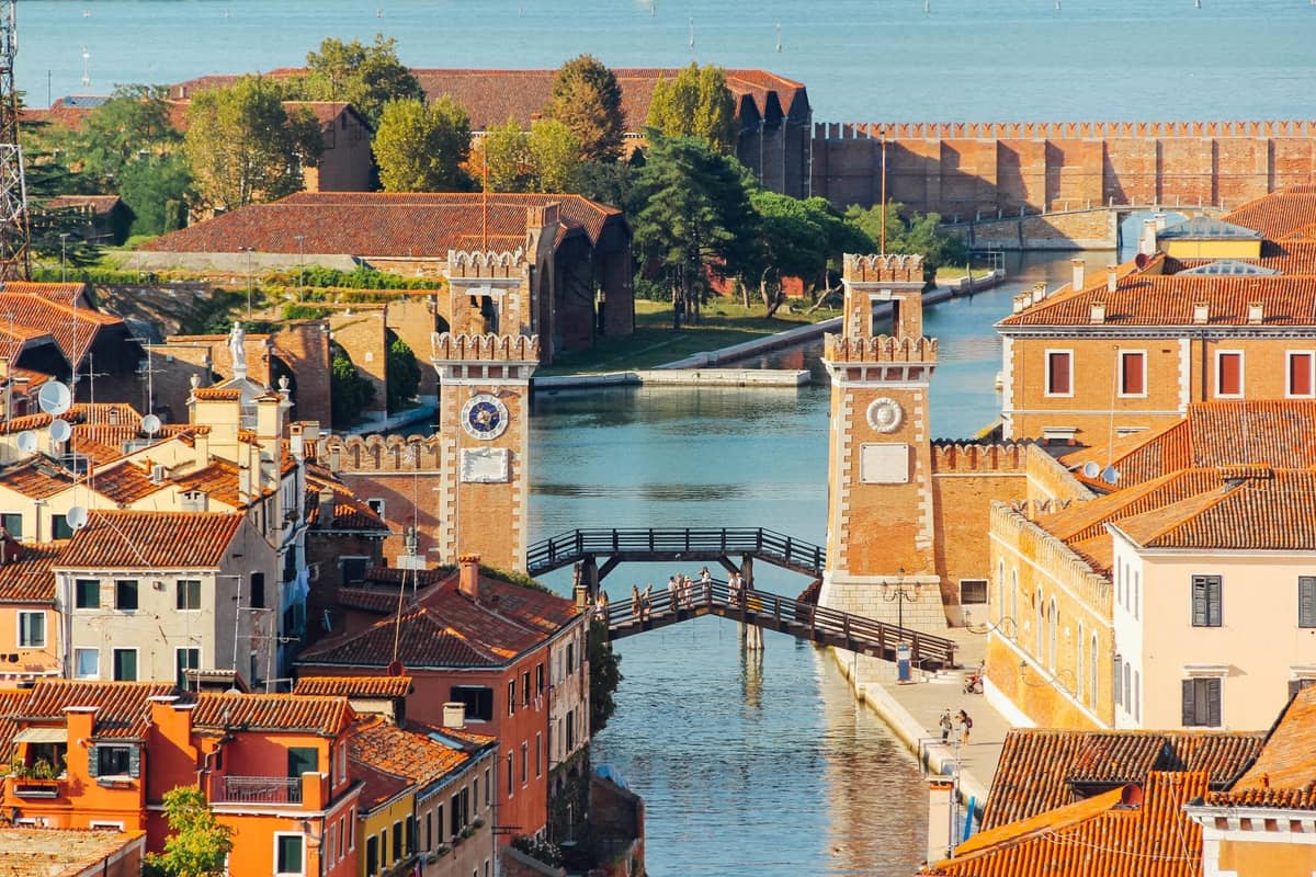 Luftaufnahme des Eingangs zum Arsenale di Venezia mit den beiden Uhrtürmen, der Holzbrücke, dem Kanal und den historischen Mauern inmitten der umliegenden Bebauung.