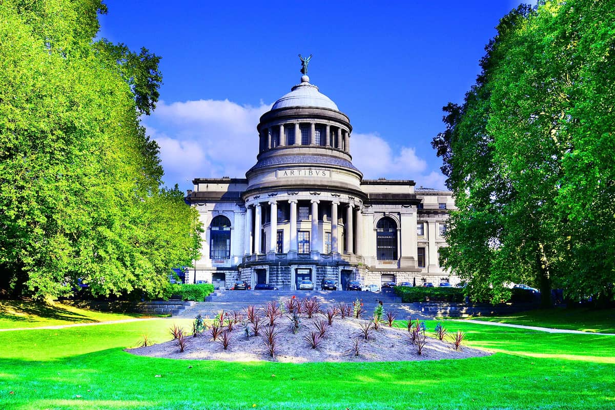 Vista frontal iluminada da cúpula neoclássica e da entrada com colunas do Museu de Arte e História, cercado por árvores frondosas no Parc du Cinquantenaire, em Bruxelas.
