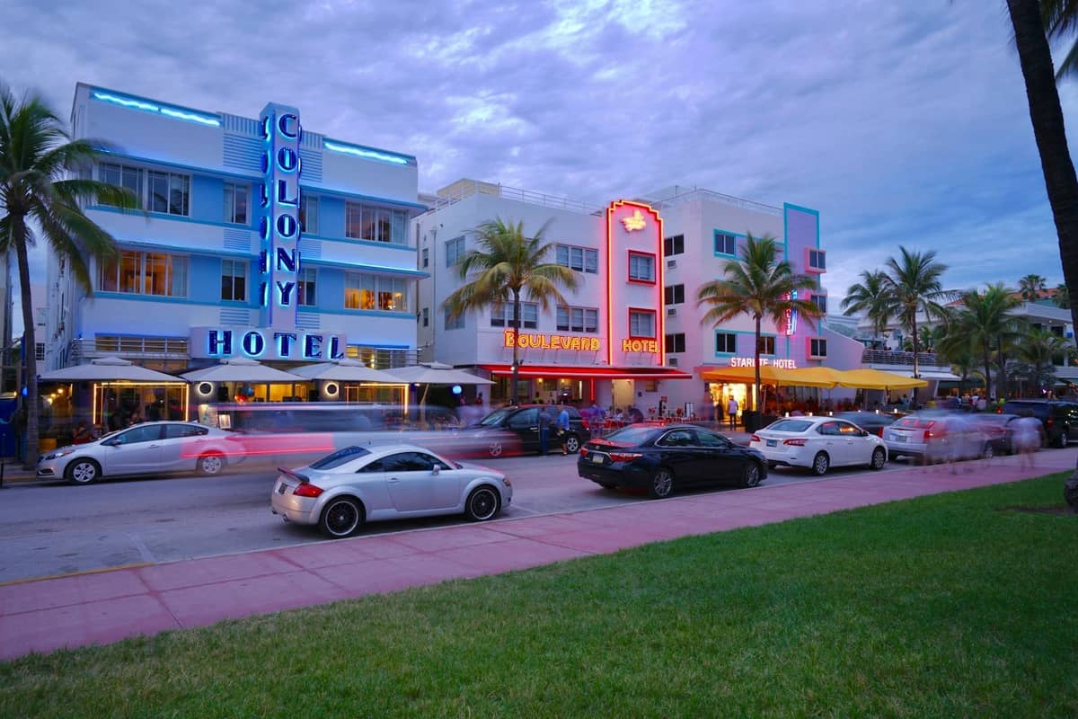 Classic pastel-colored Art Deco hotels with neon signs on Ocean Drive in Miami Beach, seen at twilight with cars and palm trees in the foreground.
