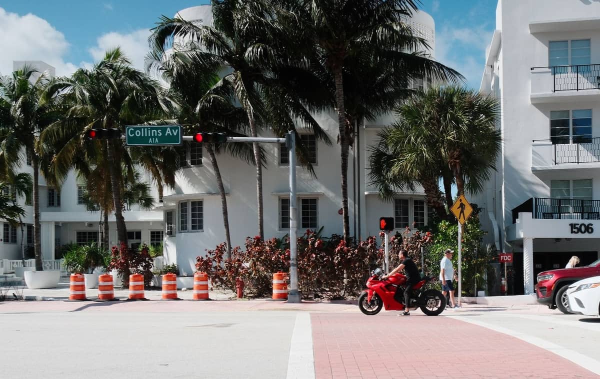 Street scene in Miami Beach with Collins Ave street sign, palm trees, people crossing, and Art Deco buildings in the background.