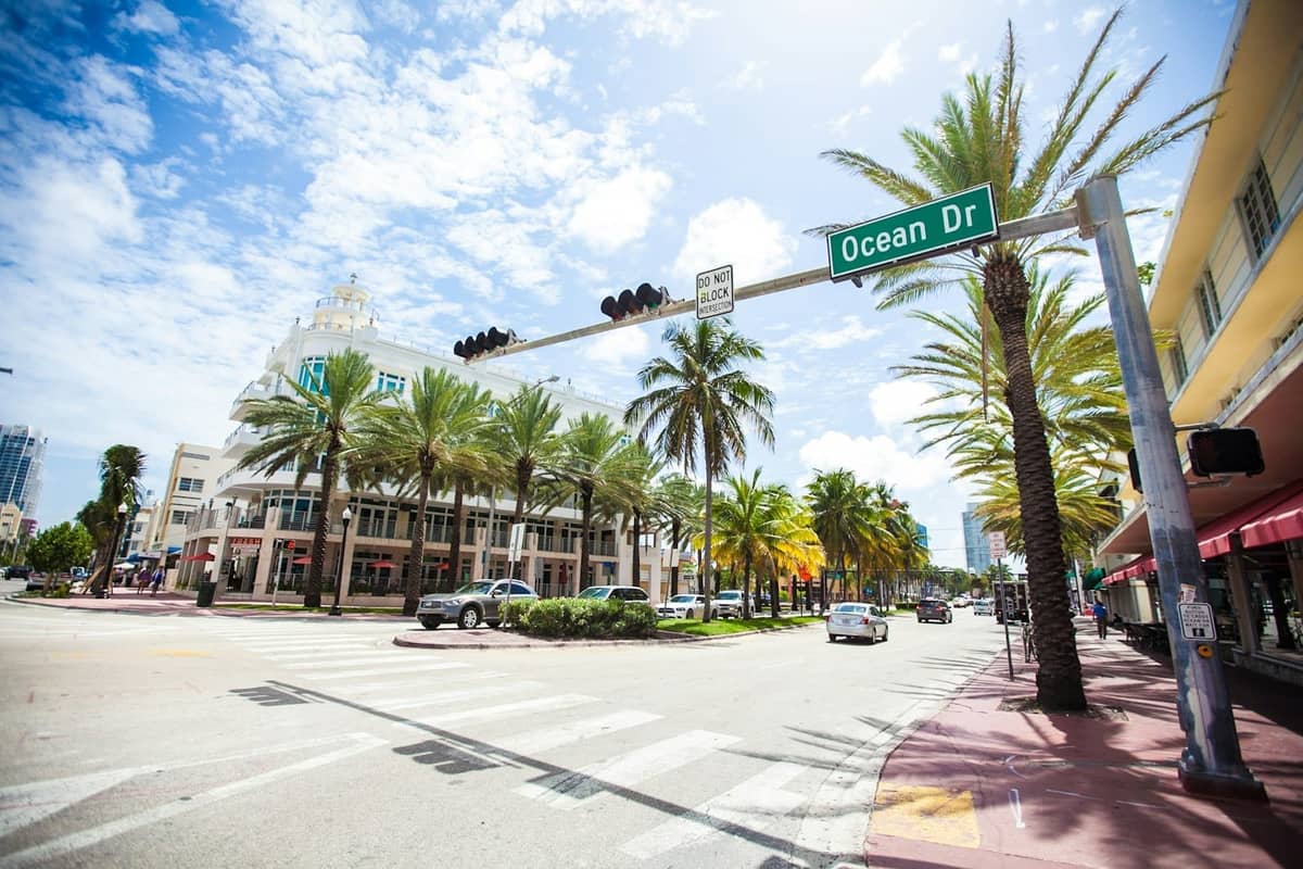Wide street view of Ocean Drive in Miami Beach with palm trees, classic Art Deco buildings, and a prominent street sign on a sunny day.
