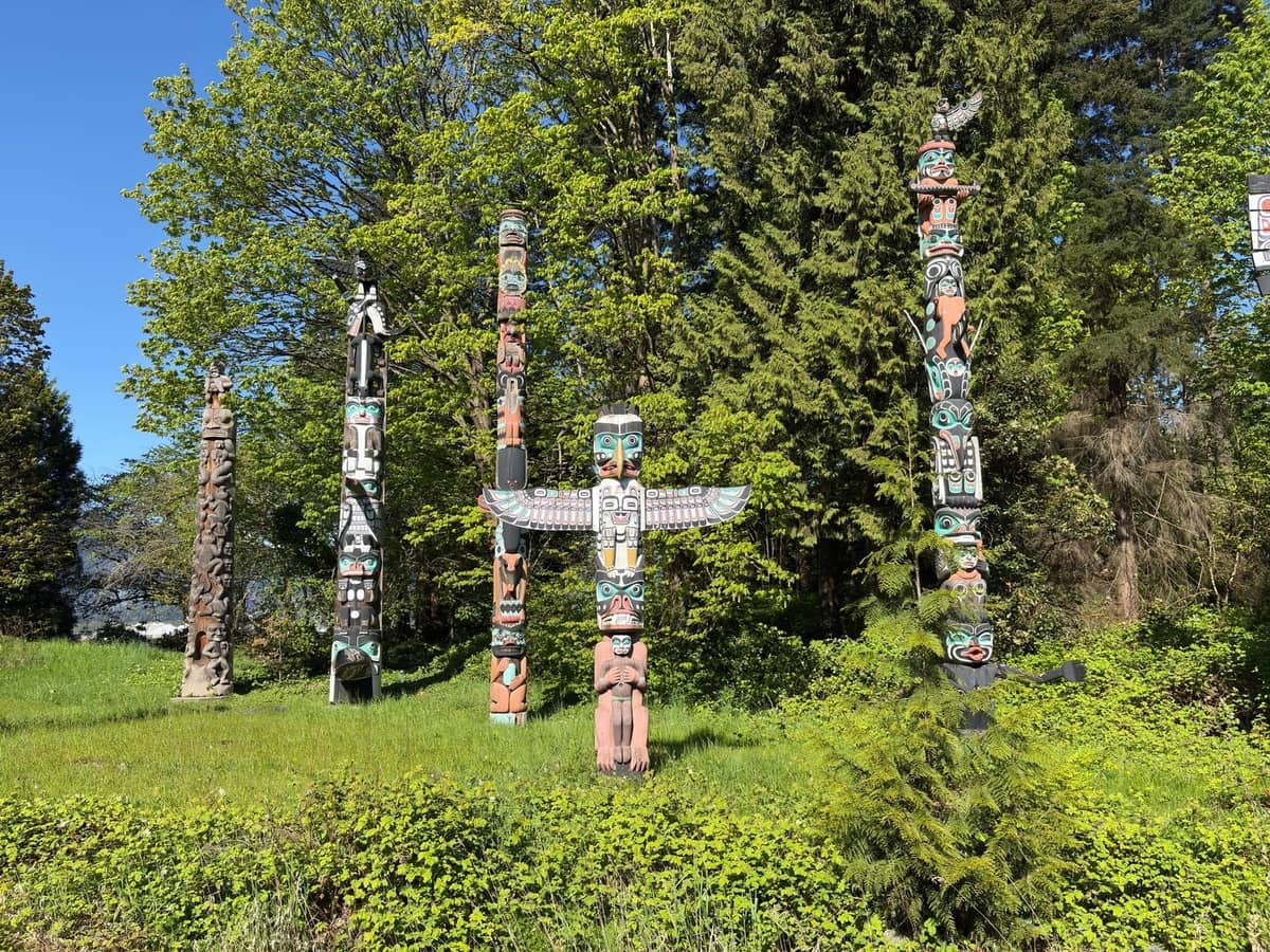 Eine Gruppe traditioneller indigener Totempfähle auf grünem Gras mit Bäumen im Hintergrund, bei Tageslicht in Vancouver fotografiert.