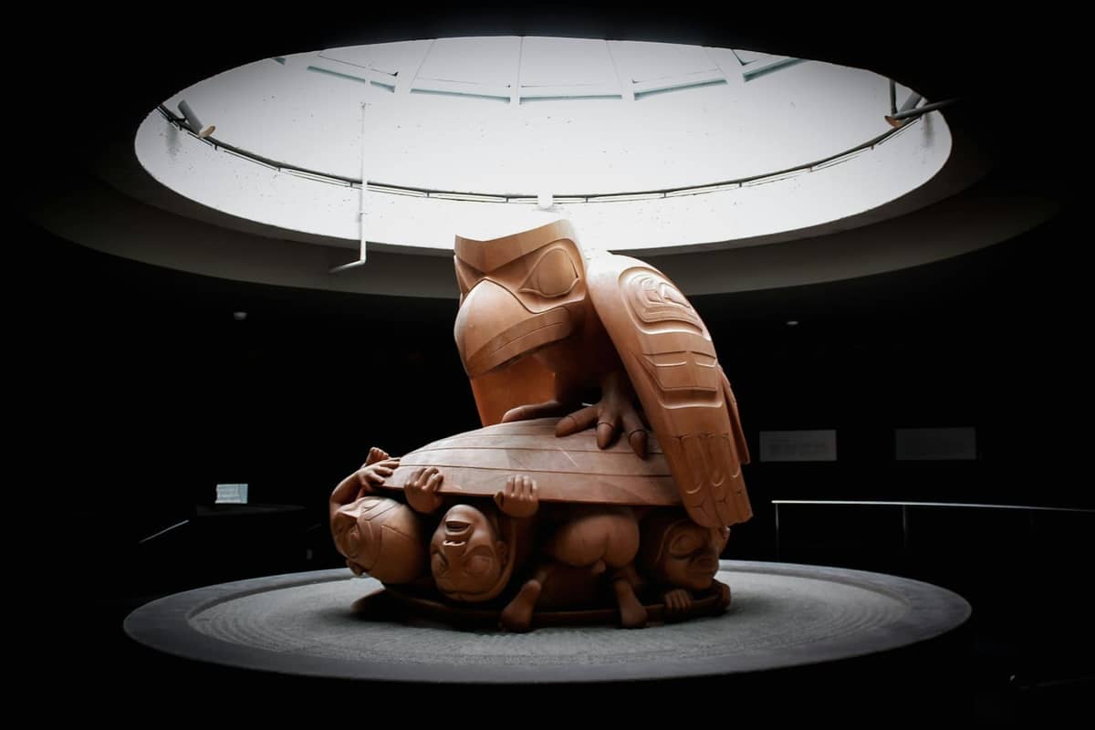 Indigene Skulptur im Innenraum mit einem Raben und menschlichen Figuren unter einem Oberlicht, ausgestellt in einem renommierten Museum mit moderner Architektur.