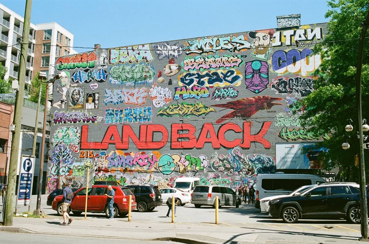 Großes Outdoor-Wandgemälde mit buntem Graffiti und dem auffälligen Schriftzug 'Land Back' an einer Wand in einem städtischen Viertel von Vancouver, mit geparkten Autos und Menschen in der Nähe.