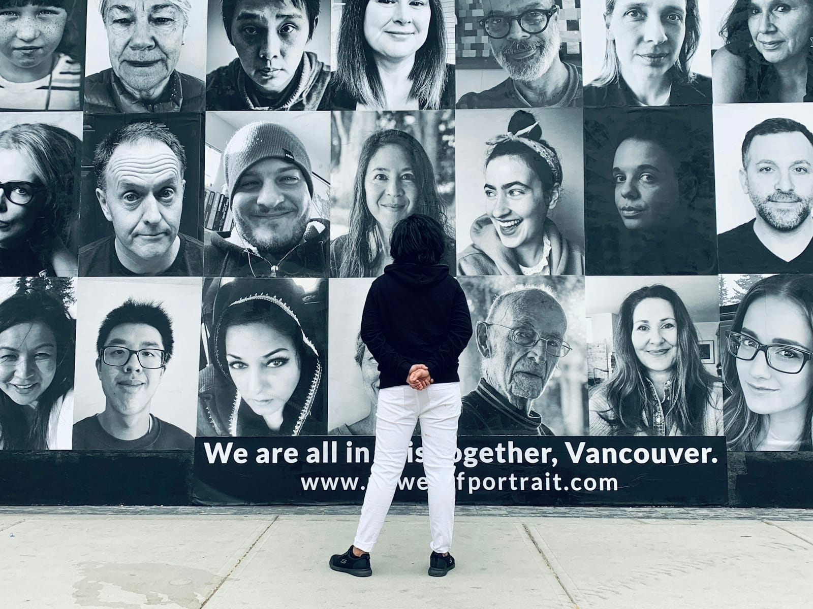 Pessoa parada diante de um grande mural externo com retratos em preto e branco de rostos diversos em Vancouver.