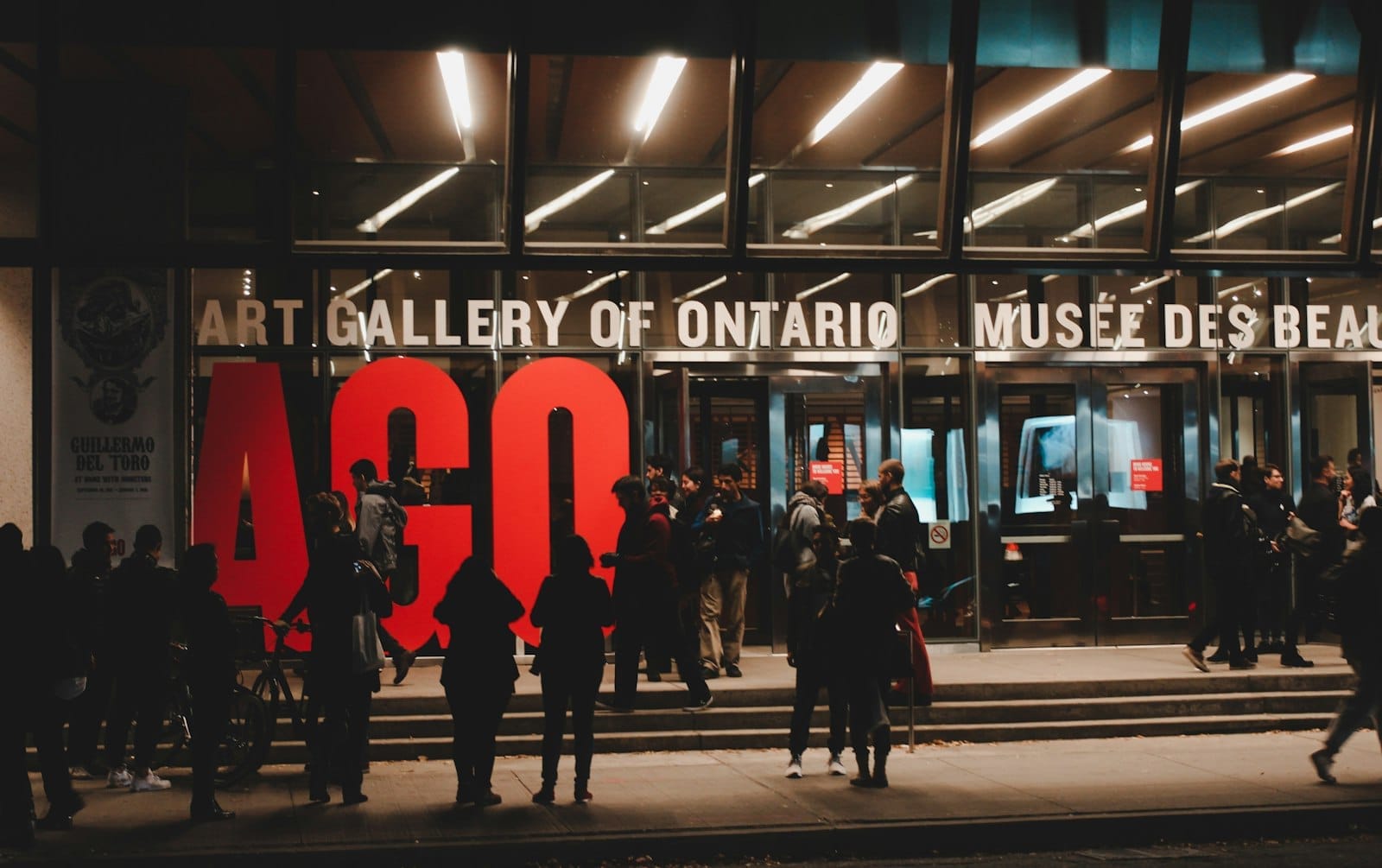 ฝูงชนยามเย็นรวมตัวกันหน้าทางเข้า Art Gallery of Ontario ที่ส่องสว่างด้วยตัวอักษร AGO สีแดงสะดุดตาและสถาปัตยกรรมกระจกสมัยใหม่