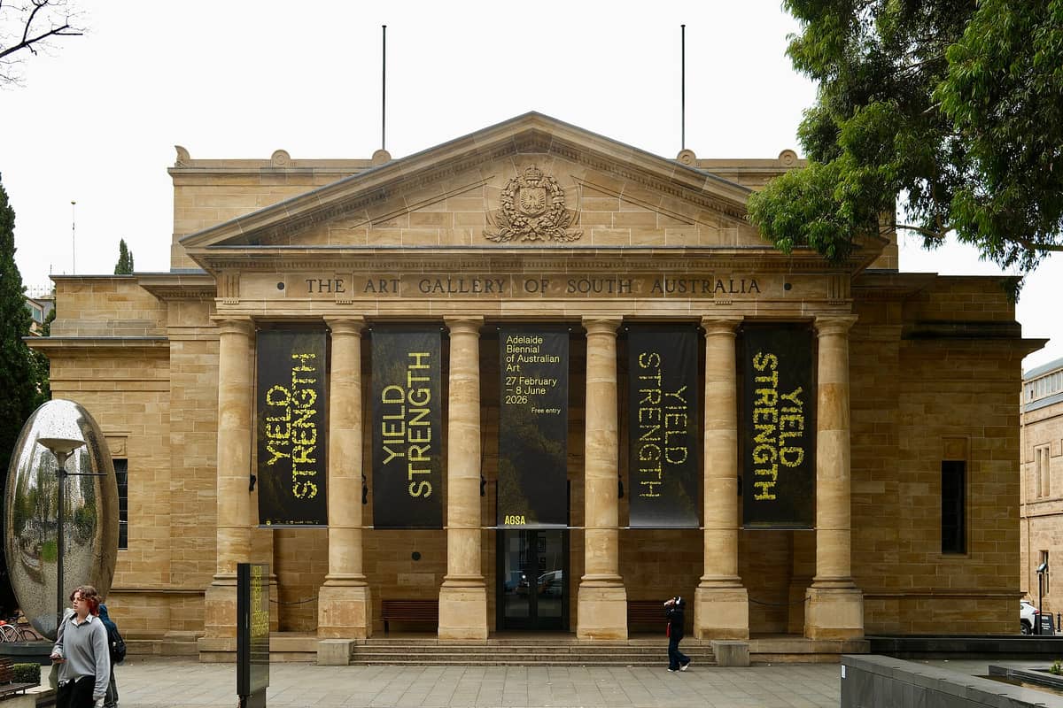 Daytime view of the Art Gallery of South Australia’s neoclassical sandstone façade with banners for the Adelaide Biennial of Australian Art and people walking outside.