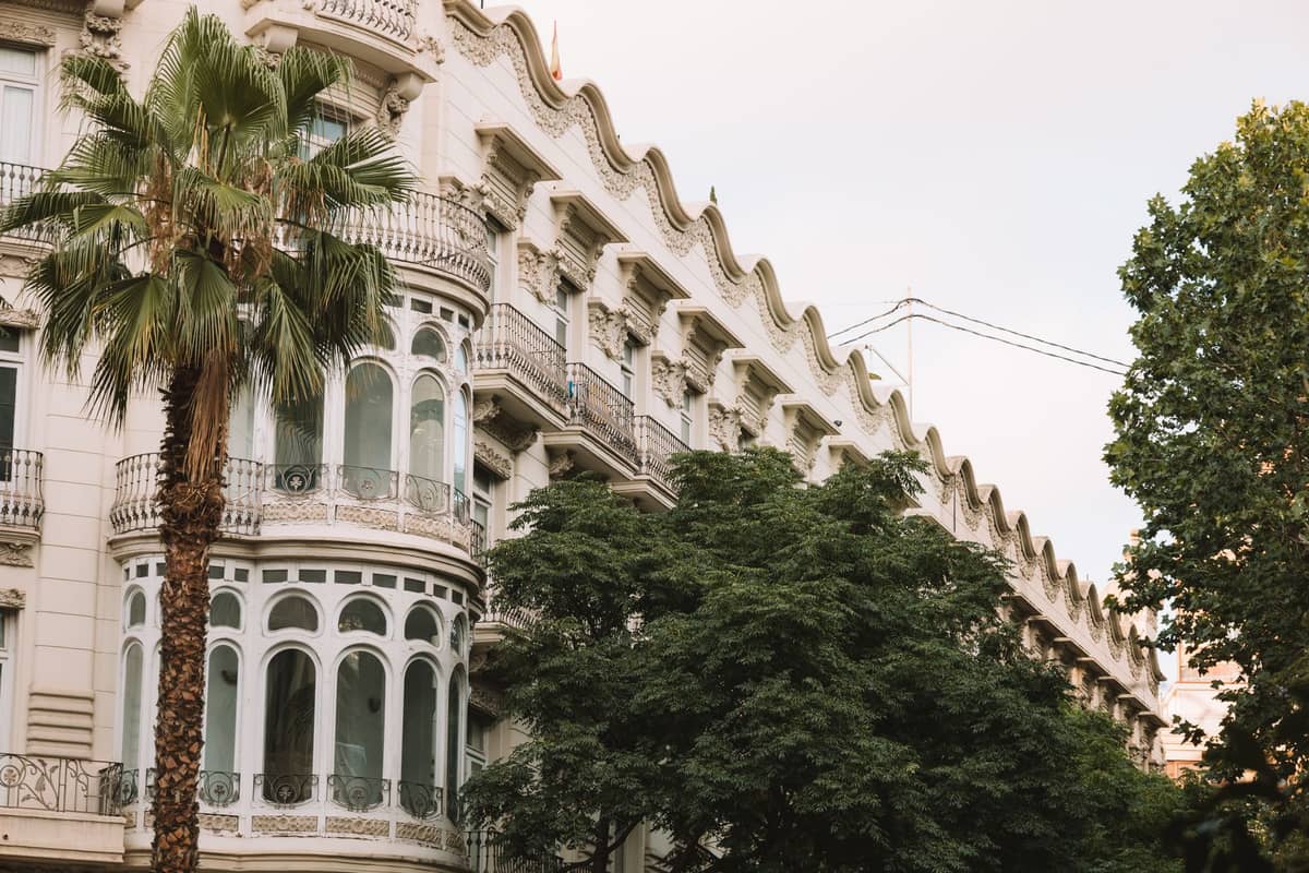 Façade d'un immeuble résidentiel orné à Valencia avec des détails Art Nouveau blancs, des balcons ondulés et une importante bow-window d'angle entourée de palmiers.