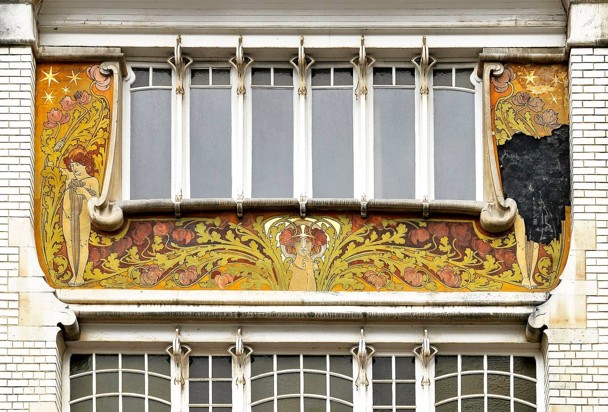 Close-up de uma fachada Art Nouveau colorida em Saint-Gilles, Bruxelas, com motivos florais decorativos e figuras femininas estilizadas ao redor de uma janela.