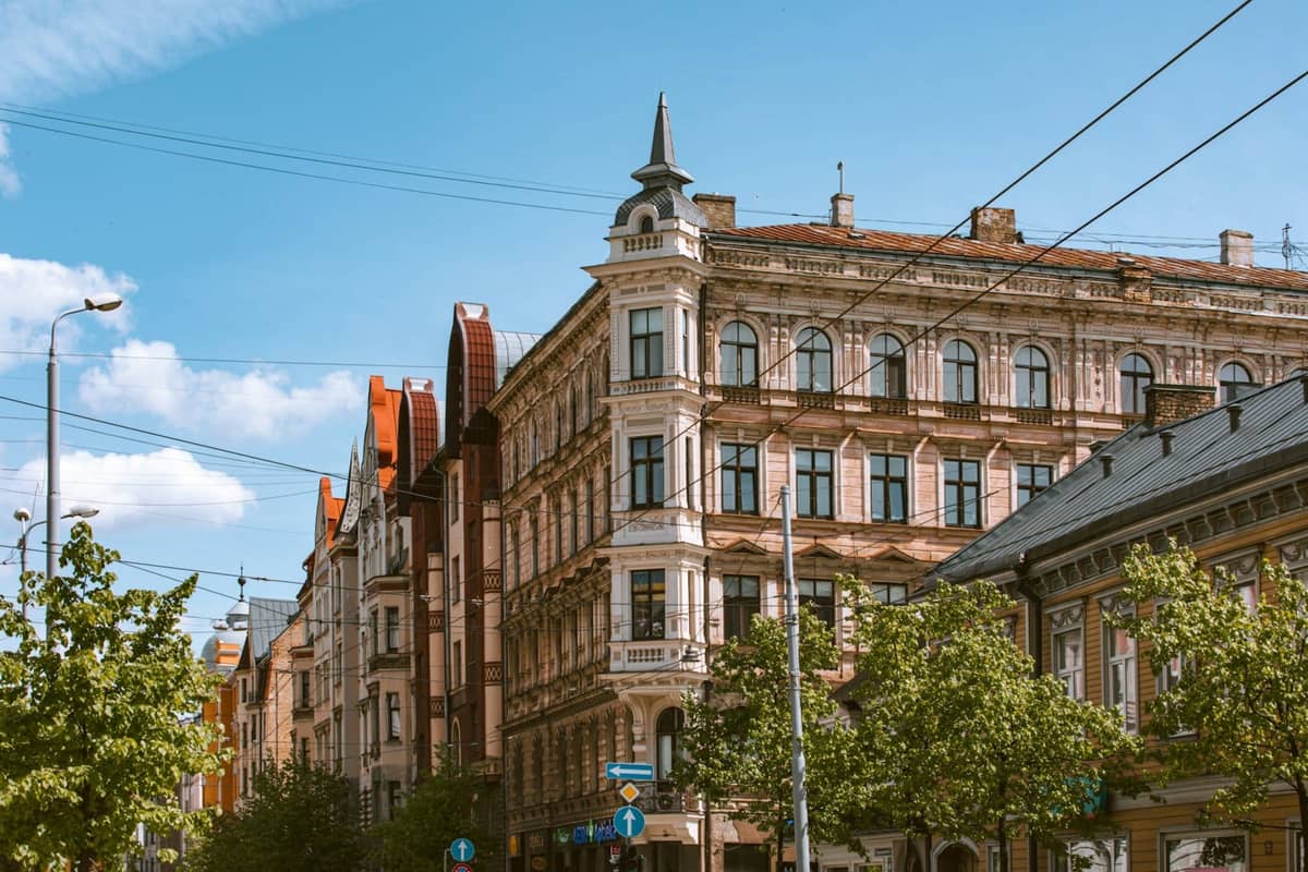 Straßenansicht von verzierten Jugendstilgebäuden in Riga mit dekorativen Fassaden und Türmen, Bäumen entlang des Bürgersteigs und Straßenbahnkabeln darüber.