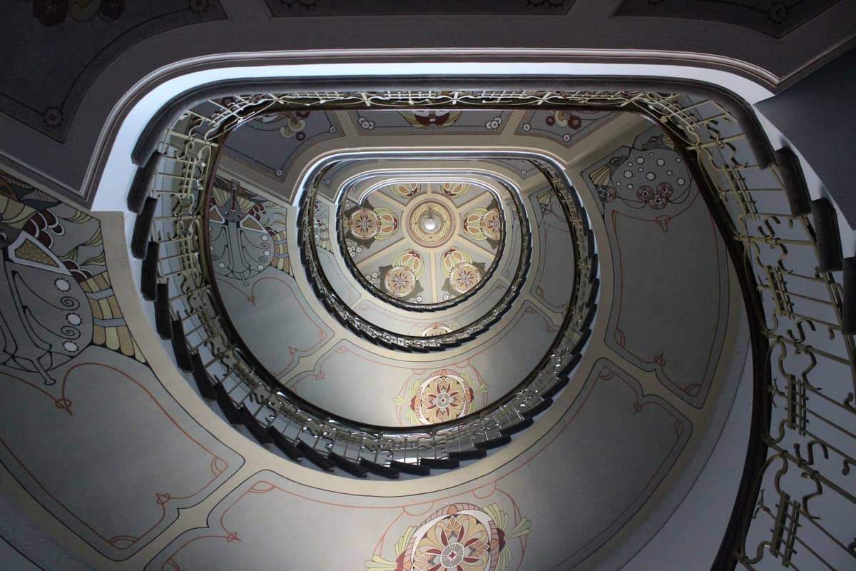 Blick nach oben auf eine spiralförmige Jugendstil-Treppe mit kunstvollen Blumenmustern und sanften Farben, die die Eleganz der historischen Innenarchitektur zeigt.