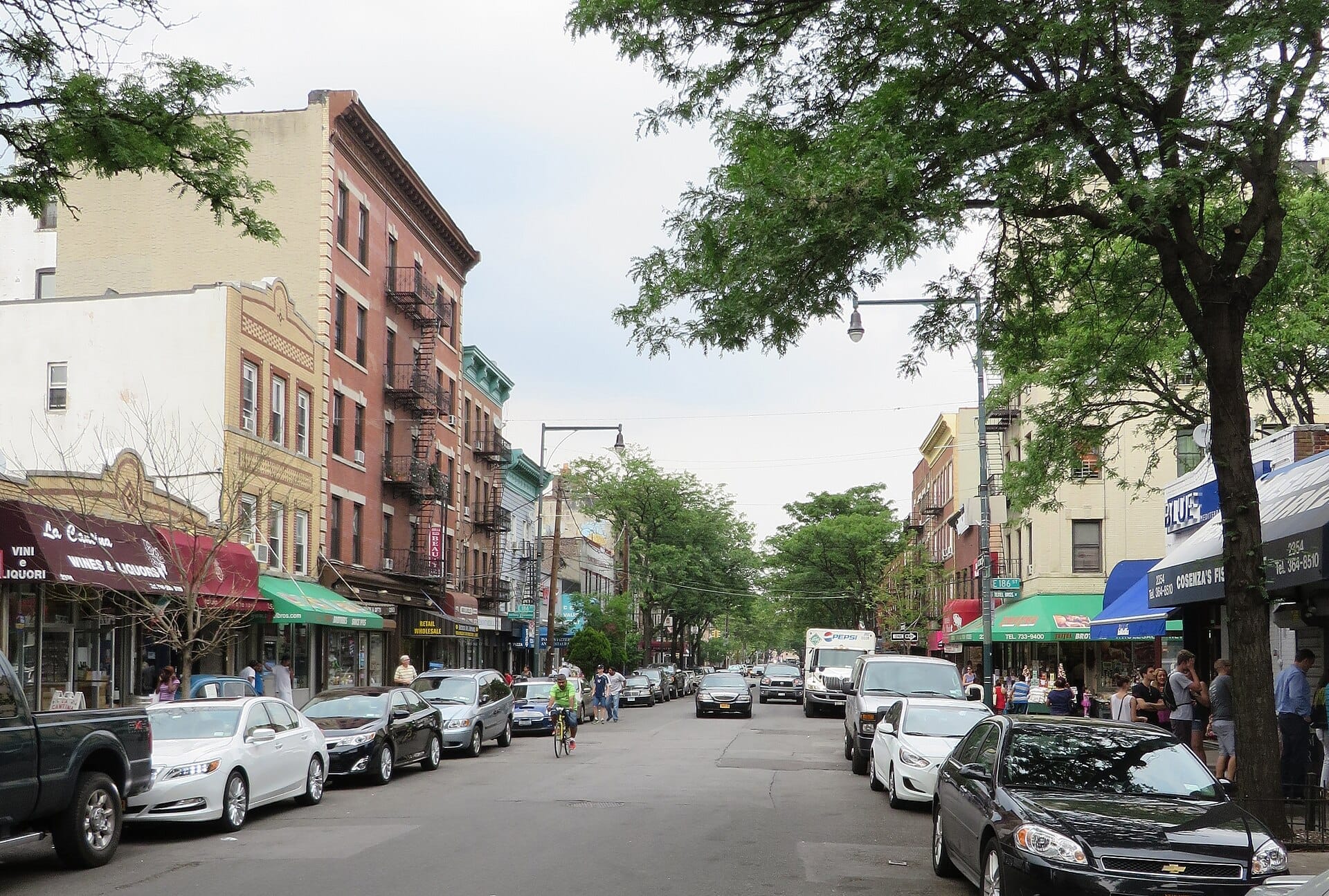 Vista stradale di Arthur Avenue nel Bronx, con storici edifici in mattoni, auto parcheggiate, tendoni di negozi e alberi frondosi in una giornata luminosa.