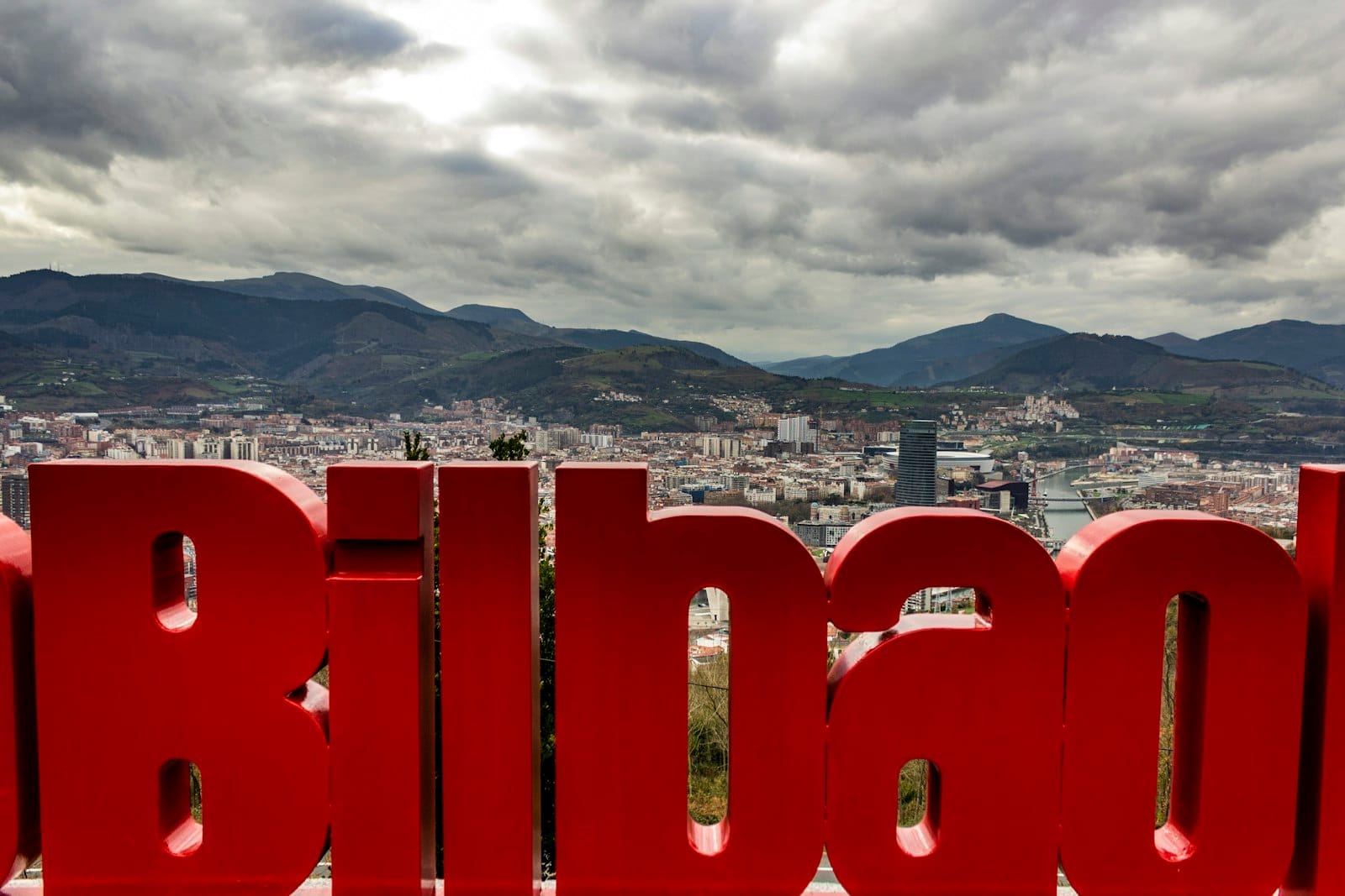 View from Artxanda Viewpoint showing Bilbao cityscape, red Bilbao sign in foreground, river, modern buildings, green hills, and dramatic cloudy sky.