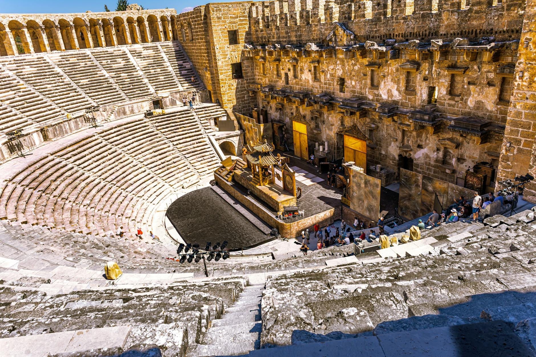 Vista grandangolare del Teatro Antico di Aspendos con il palcoscenico allestito per lo spettacolo, la platea gremita e una calda luce dorata che illumina l'anfiteatro romano splendidamente conservato.