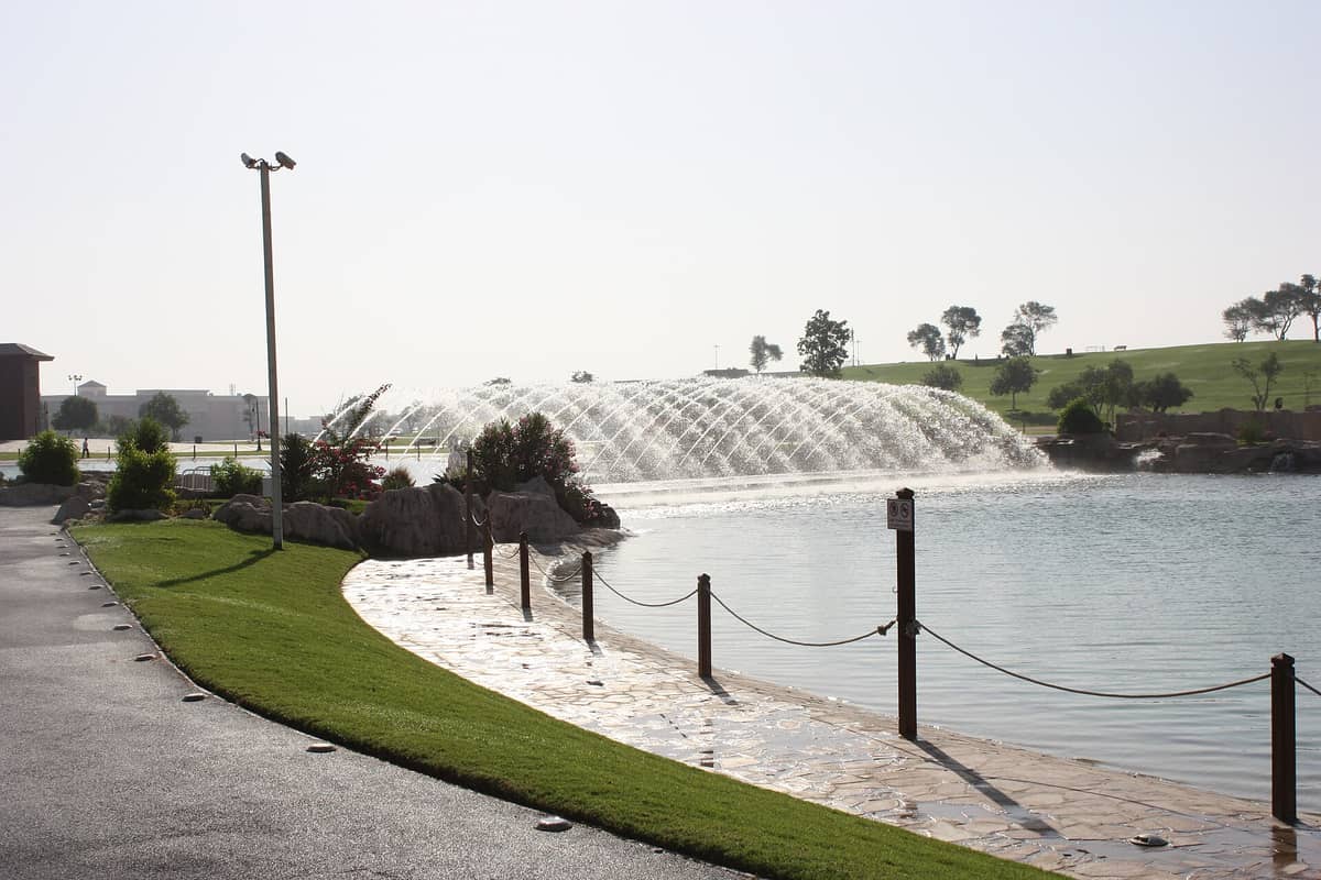 Der Seeweg im Aspire Park mit einem großen dekorativen Wasserfontäne über dem Wasser, grünen Rasenflächen und Bäumen im Hintergrund an einem sonnigen Tag.