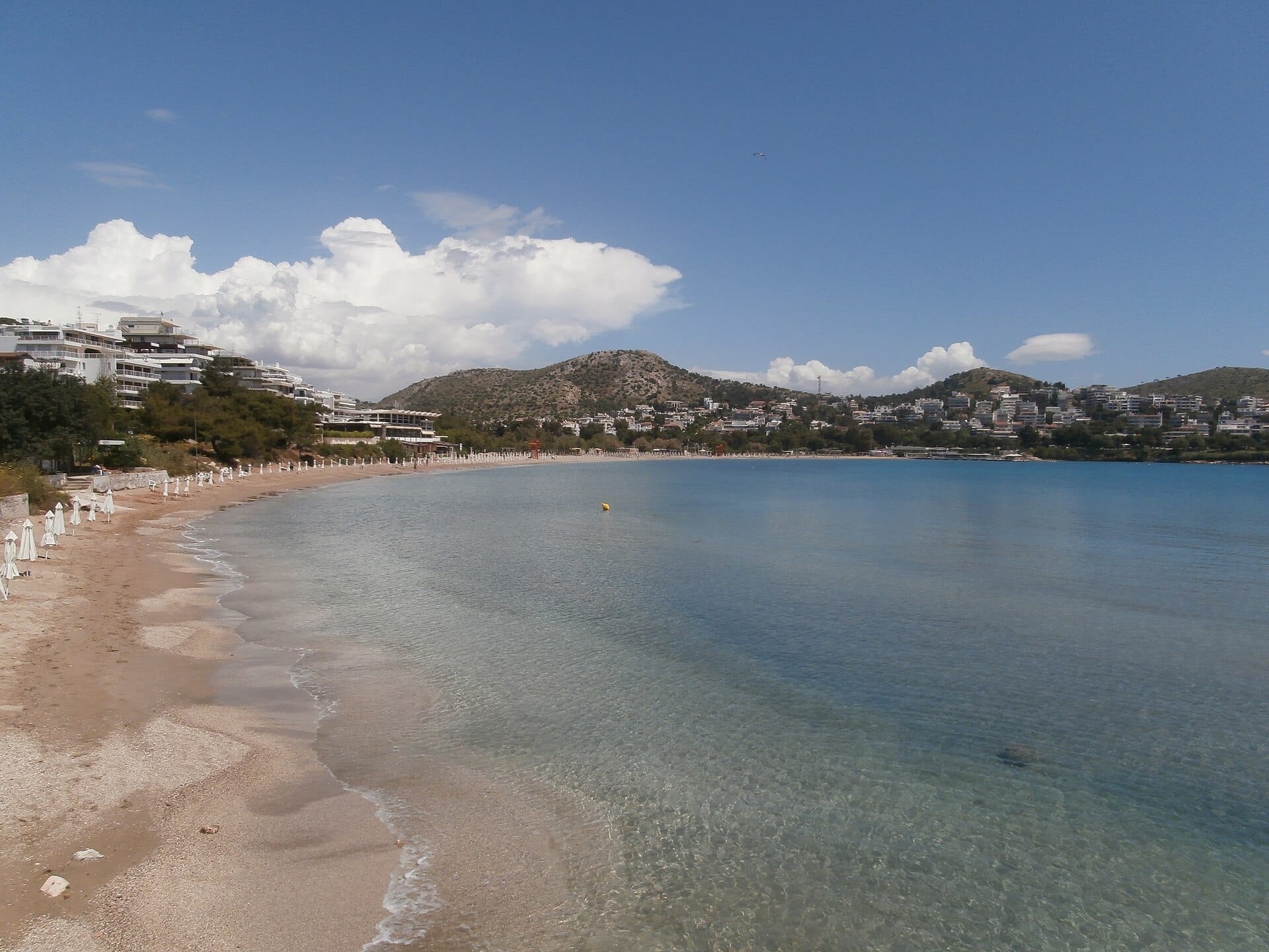 ชายหาดทรายกว้างบน Athenian Riviera พร้อมน้ำทะเลสีฟ้าใส ร่มสีขาว อาคารสมัยใหม่ และเนินเขาสีเขียวภายใต้ท้องฟ้าสดใส
