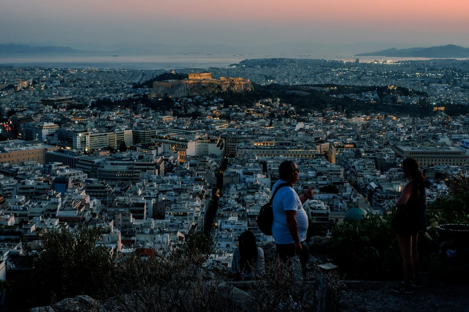 Vista da una collina ad Atene al tramonto con persone in primo piano che guardano sulla città e l'Acropoli illuminata in lontananza.