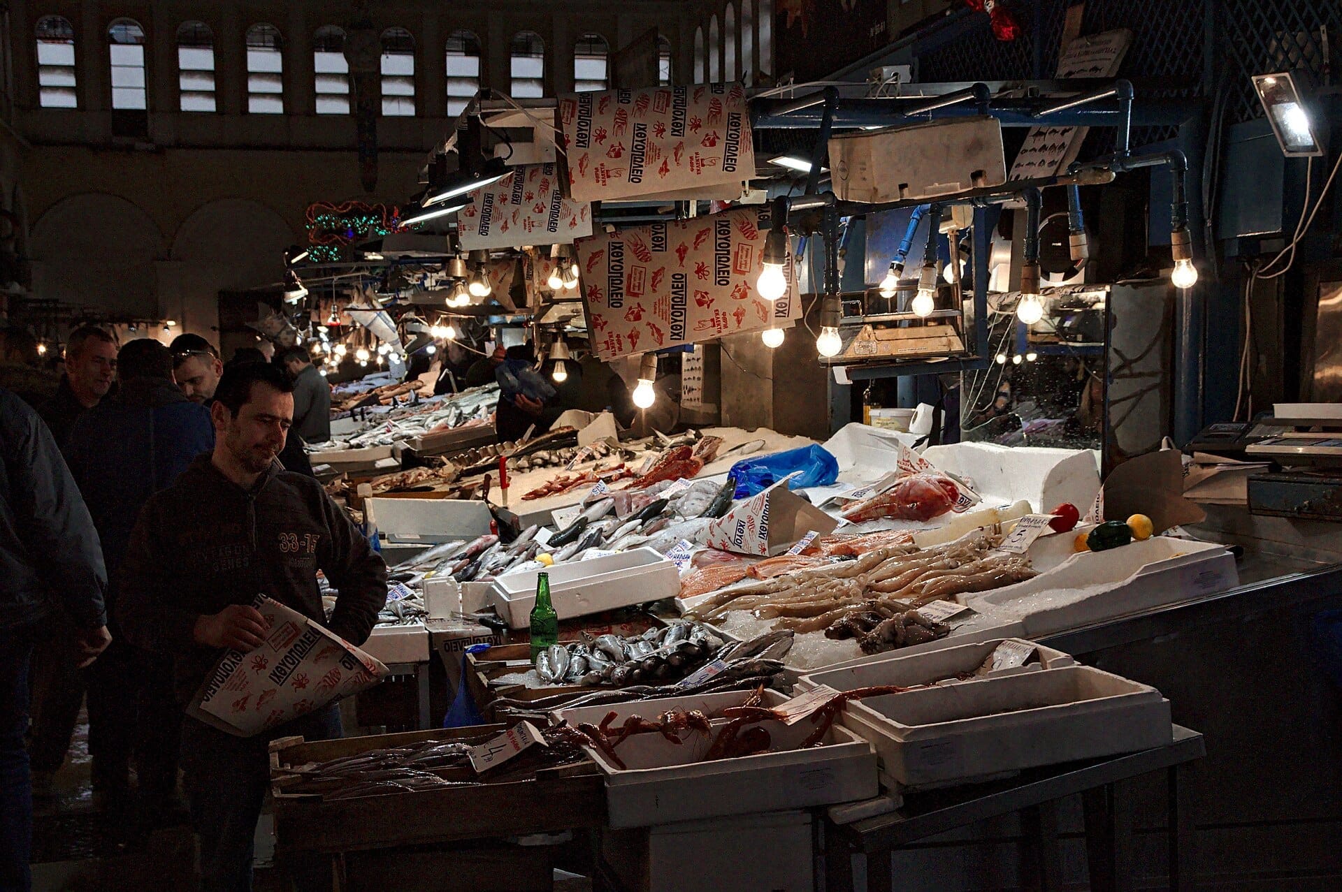 Pescivendoli e acquirenti si radunano intorno a bancarelle di frutti di mare illuminate al Mercato Centrale di Atene, con pesce fresco e polpo esposti sul ghiaccio sotto lampadine appese.