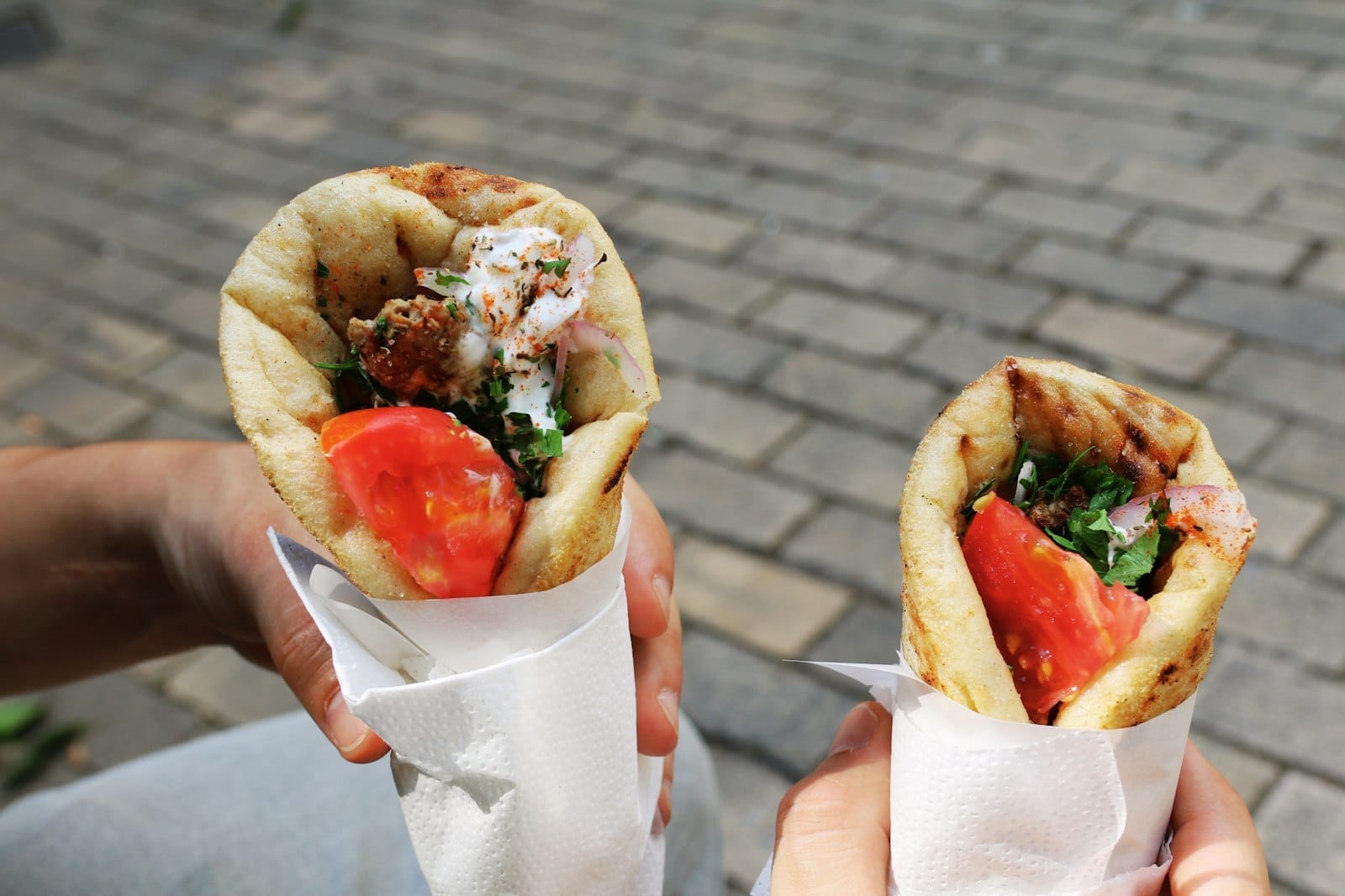 Duas mãos segurando um gyros grego fresquinho recheado com carne, tomate, ervas e molho, numa rua de pedras ensolarada em Atenas.