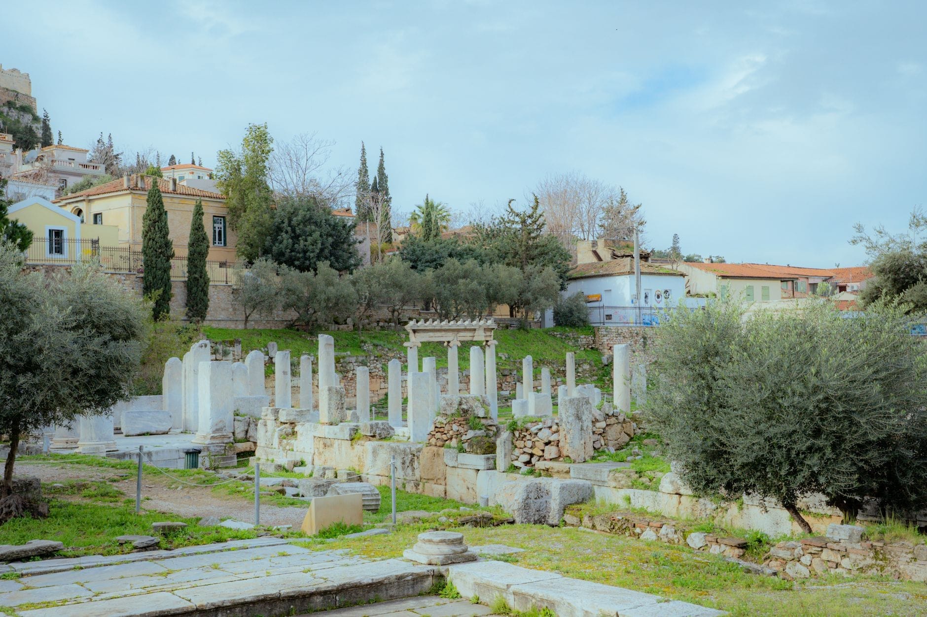 Starożytne kamienne ruiny i stojące kolumny otoczone zielenią na terenie stanowiska archeologicznego z domami i drzewami w tle.
