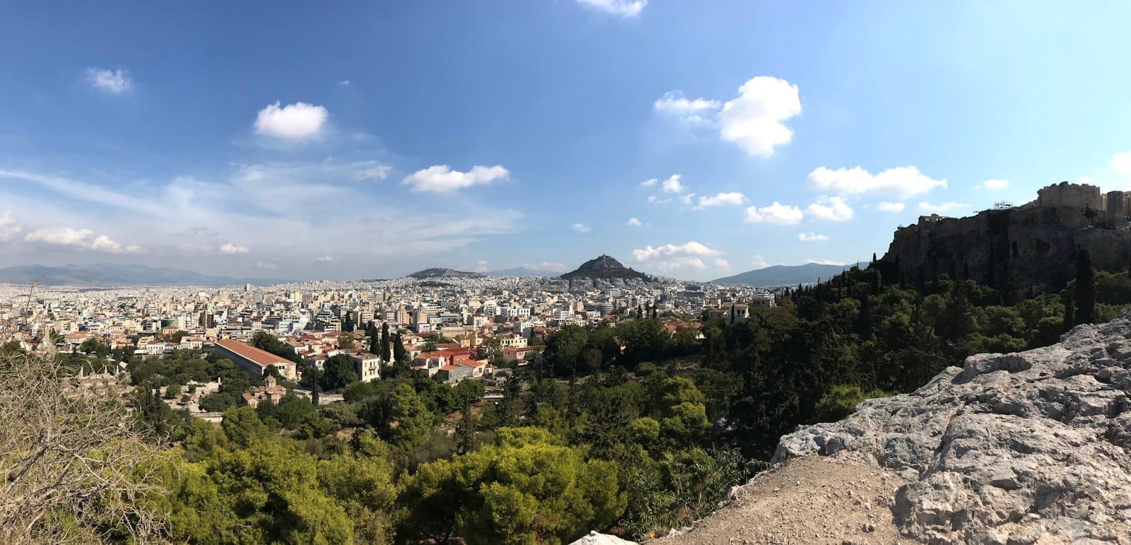Vista panorâmica de Atenas a partir de uma colina, com prédios, árvores verdes, morros ao fundo e céu azul com nuvens esparsas. Uma formação rochosa emoldura o primeiro plano.