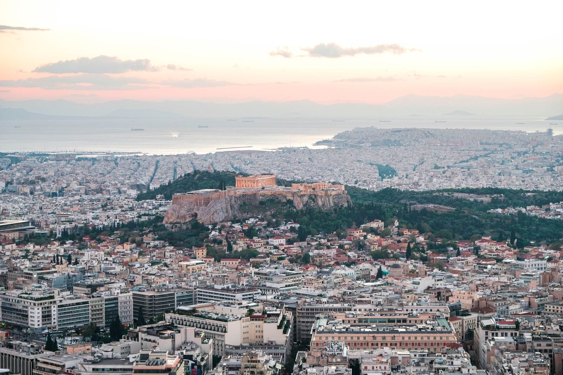Vista panorâmica de Atenas ao pôr do sol com a Acrópole iluminada no alto de uma colina, a cidade se espalhando ao fundo e um suave céu rosado sobre o mar ao longe.