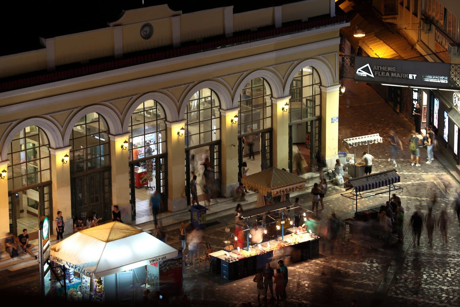 Cena noturna movimentada em Atenas, com pessoas reunidas do lado de fora de um prédio bem iluminado, barracas de mercado e ruas iluminadas, capturando a atmosfera vibrante da cidade à noite.
