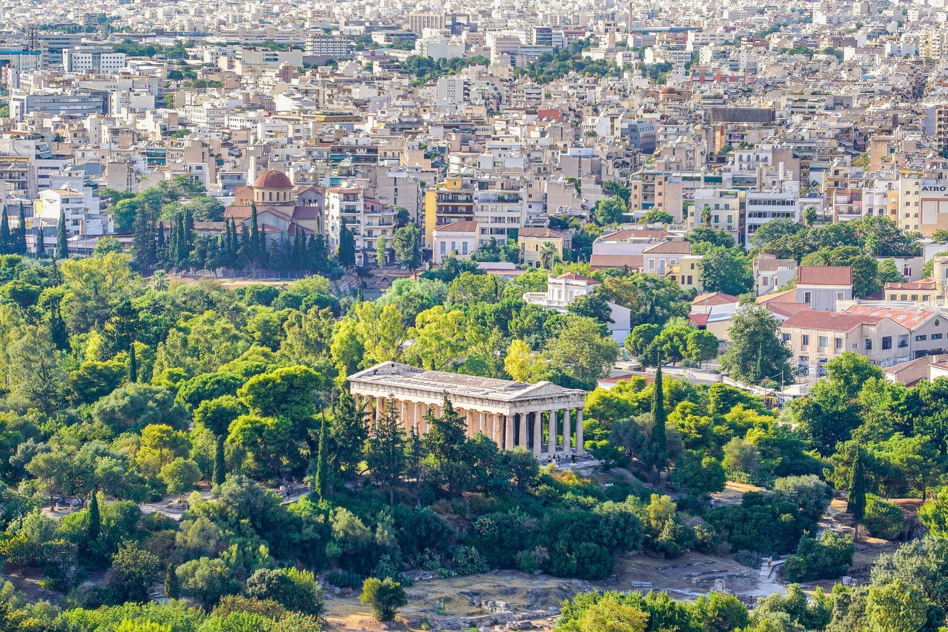 Vista aerea di Atene con l'antico Tempio di Efesto circondato da alberi verdi e il paesaggio urbano sullo sfondo.