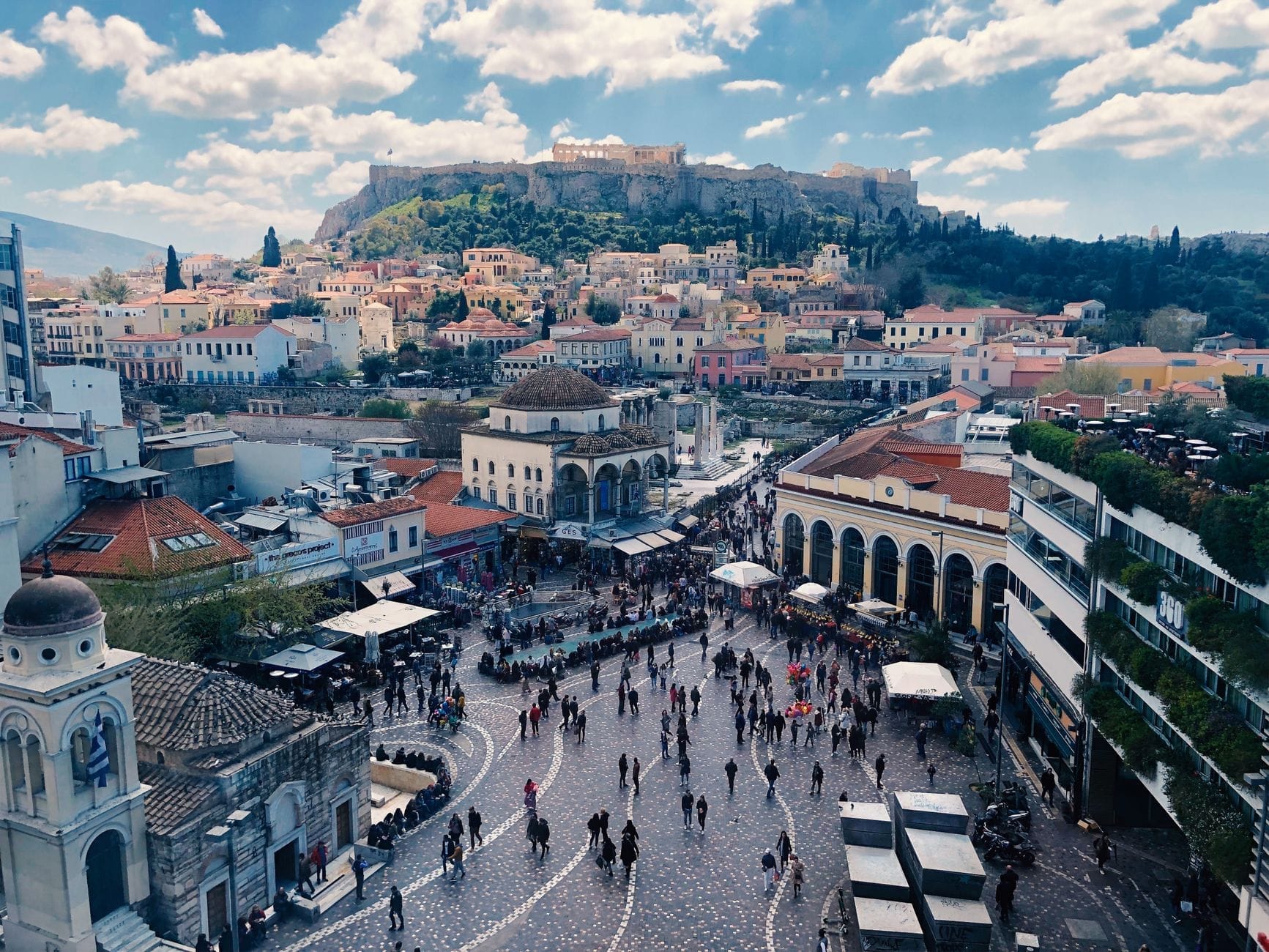 Vista aerea di Piazza Monastiraki ad Atene con l'Acropoli sullo sfondo, animata dalla folla e circondata da edifici economici.