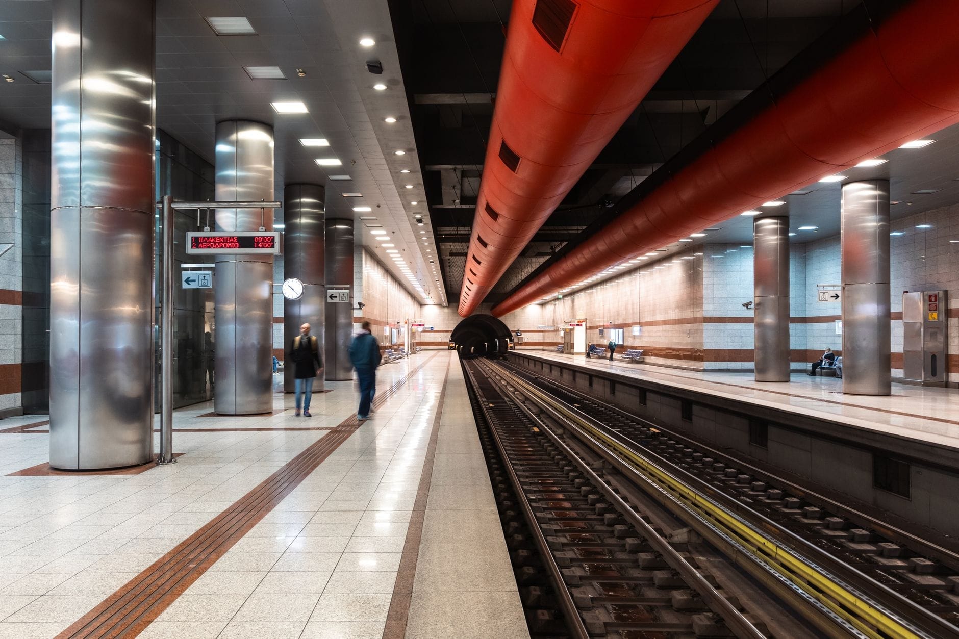 Moderna stazione della metropolitana di Athens con binari, soffitto rosso e alcune persone in attesa sulla banchina.